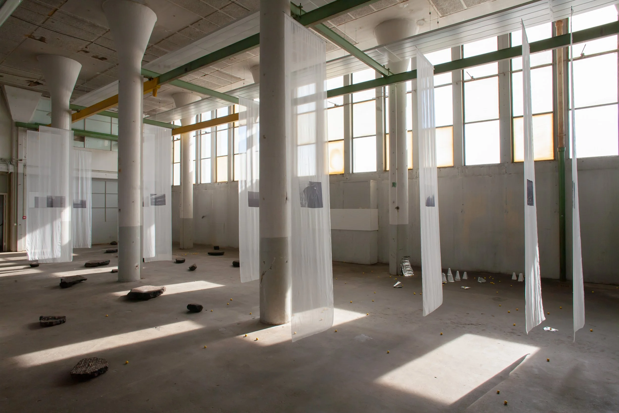 Interior of a building with white curtains hanging from a green and yellow beam. The ceiling is unfinished with visible concrete and pipes. The floor has scattered rocks and debris, with large windows on the upper wall allowing sunlight to illuminate the space.