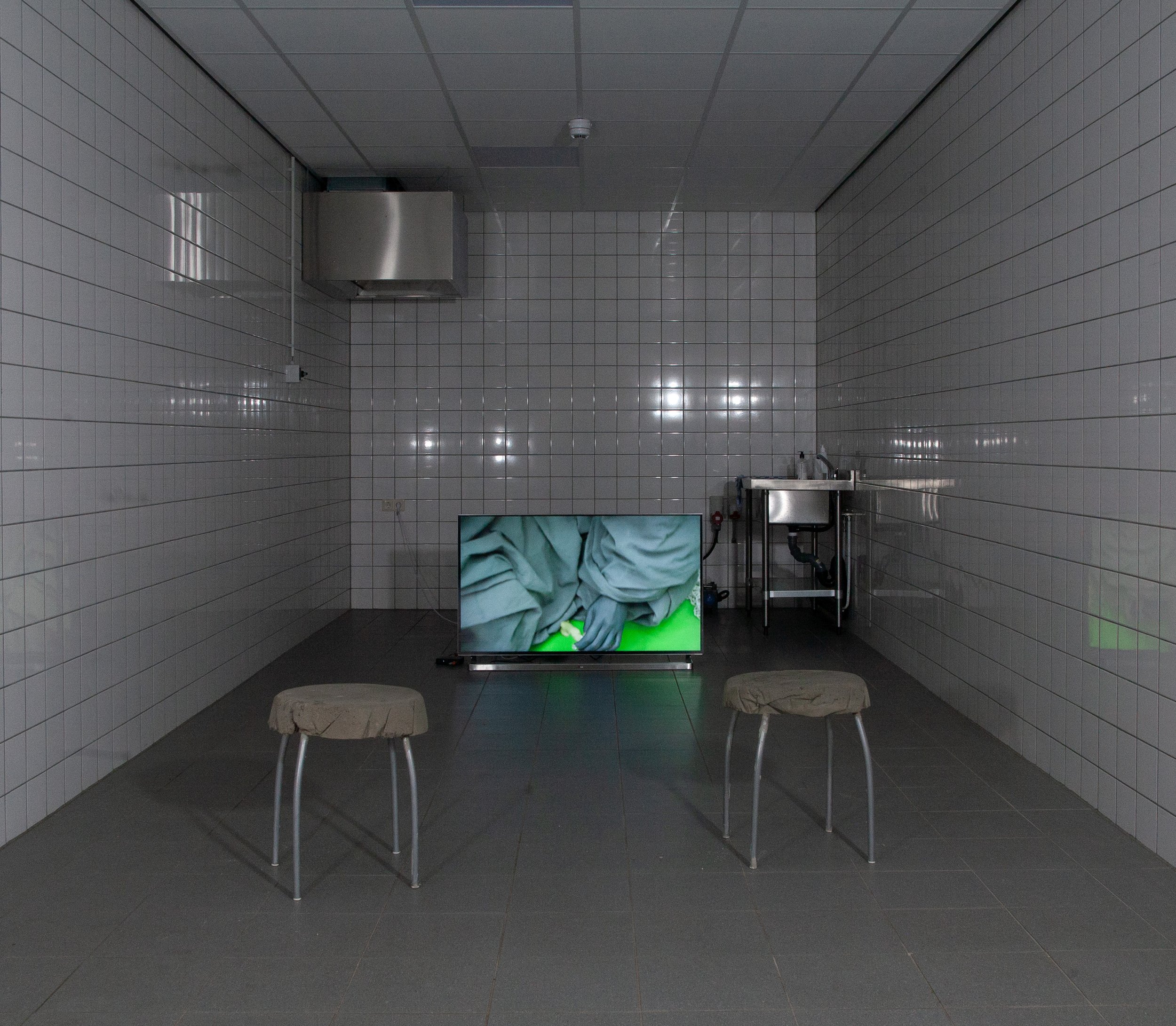 A bare, tiled room resembling a kitchen or medical facility with gray floor and walls, a stainless steel sink, stools, and a TV screen displaying an image of a person in scrubs.