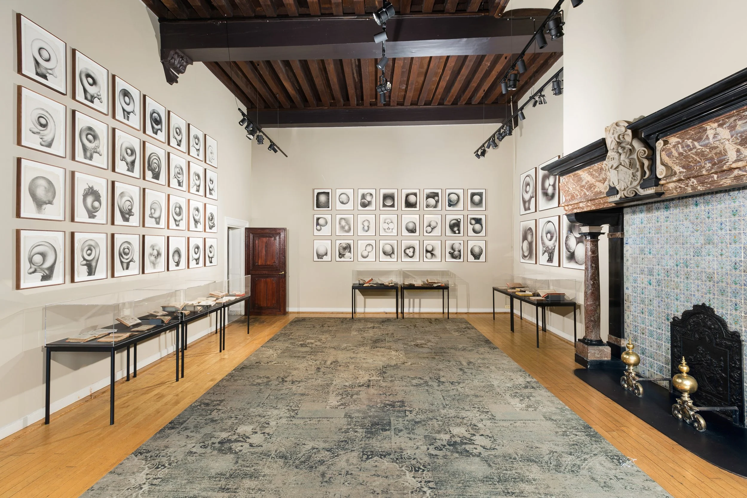 Art gallery displaying black and white framed sketches of abstract faces and heads on cream walls, with display tables holding books, a stone fireplace with ornate carving, and a patterned tile section on the right wall, wooden ceiling, and hardwood flooring.
