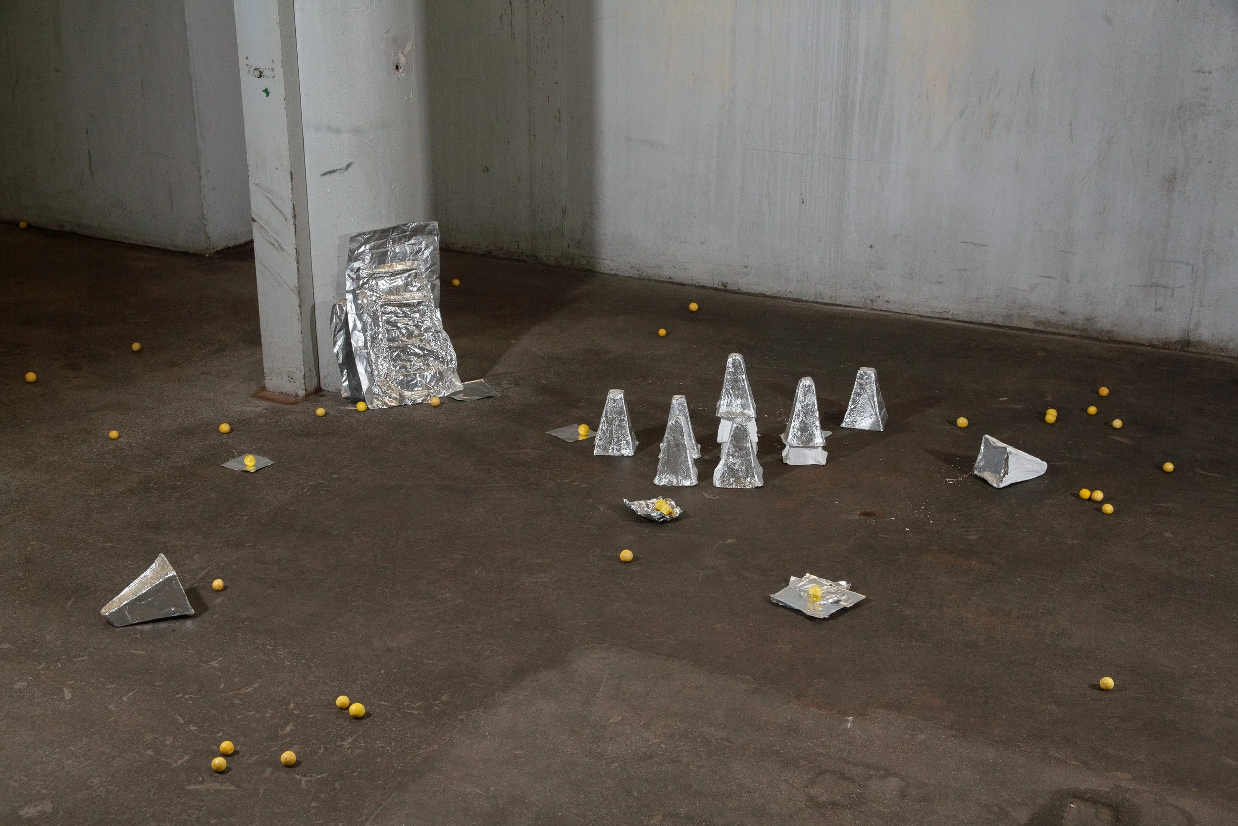 A concrete wall and floor scattered with yellow ping pong balls, broken pieces of foil, and crumpled foil wrappers.