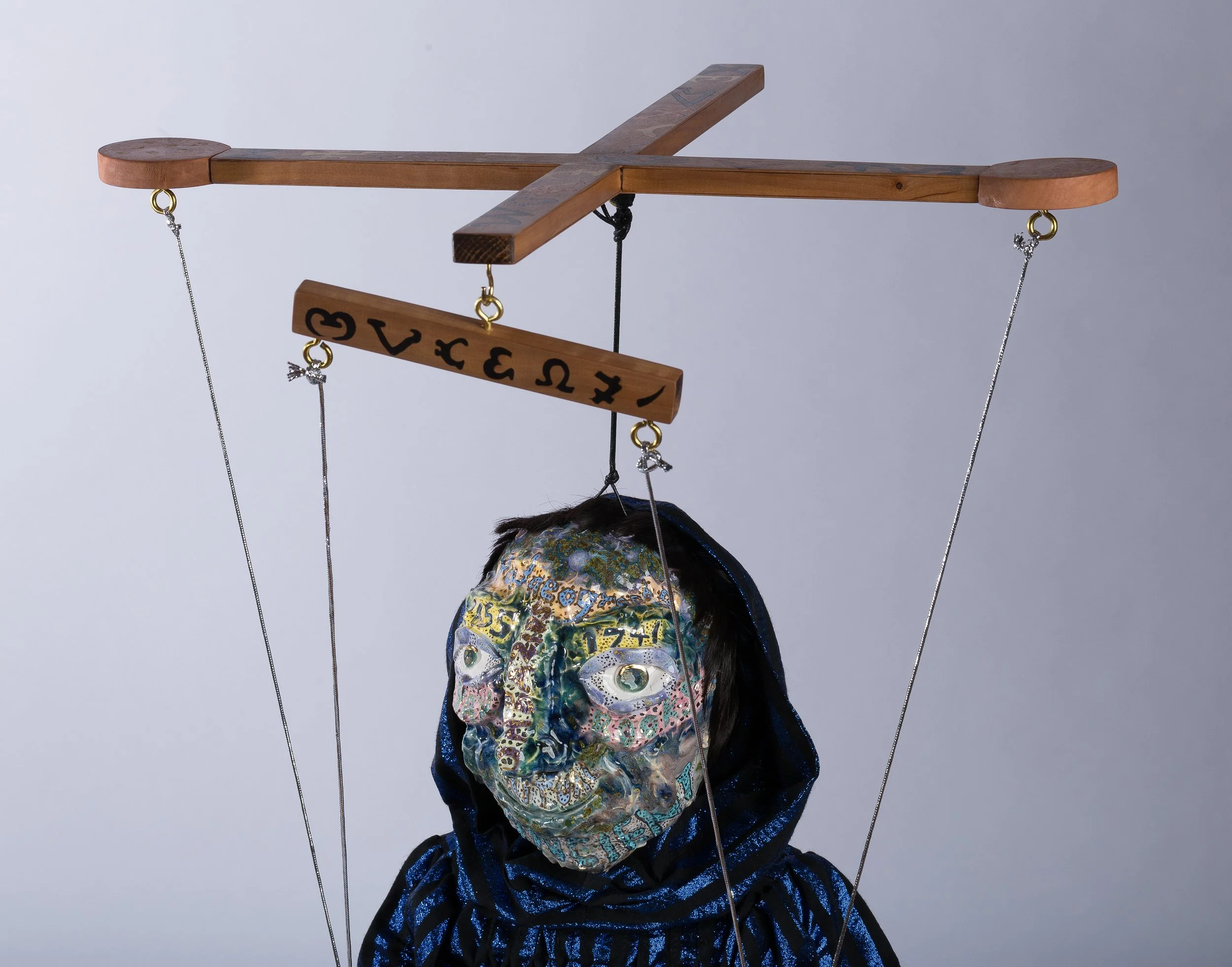 A puppet with a large, colorful, textured face and a black velvet body, hanging from a wooden cross-shaped structure with strings.