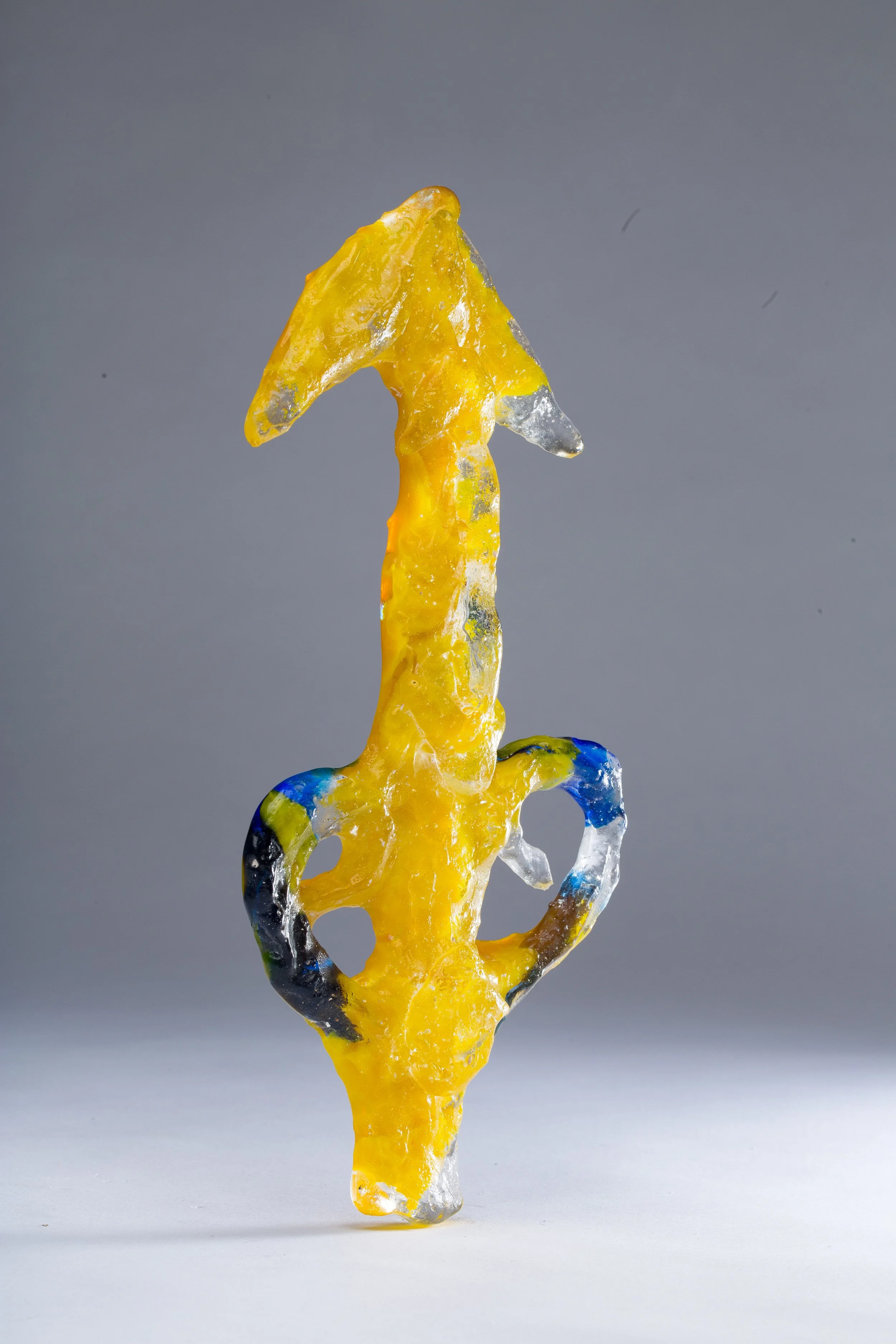 Colorful glass sculpture resembling a flame or a stylized figure with yellow, blue, black, and clear glass elements, set against a gray background.