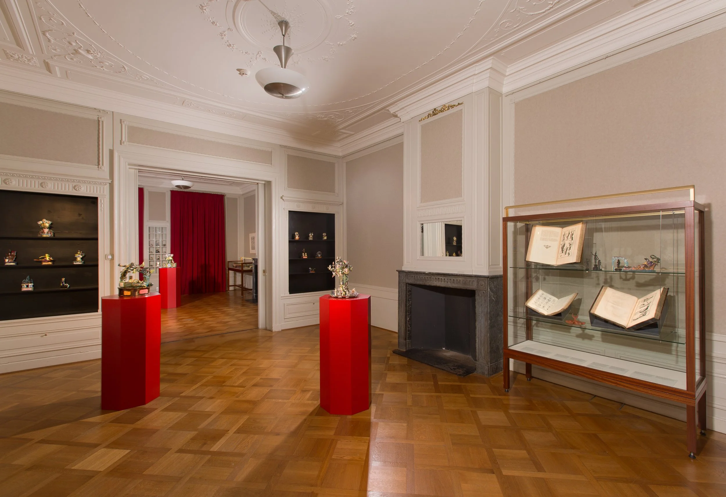 An interior of a museum or gallery room with display cases and shelves containing artifacts, decorative molding on the ceiling, a white mantel fireplace, and red pedestals with art pieces, with a doorway and red curtains in the background.