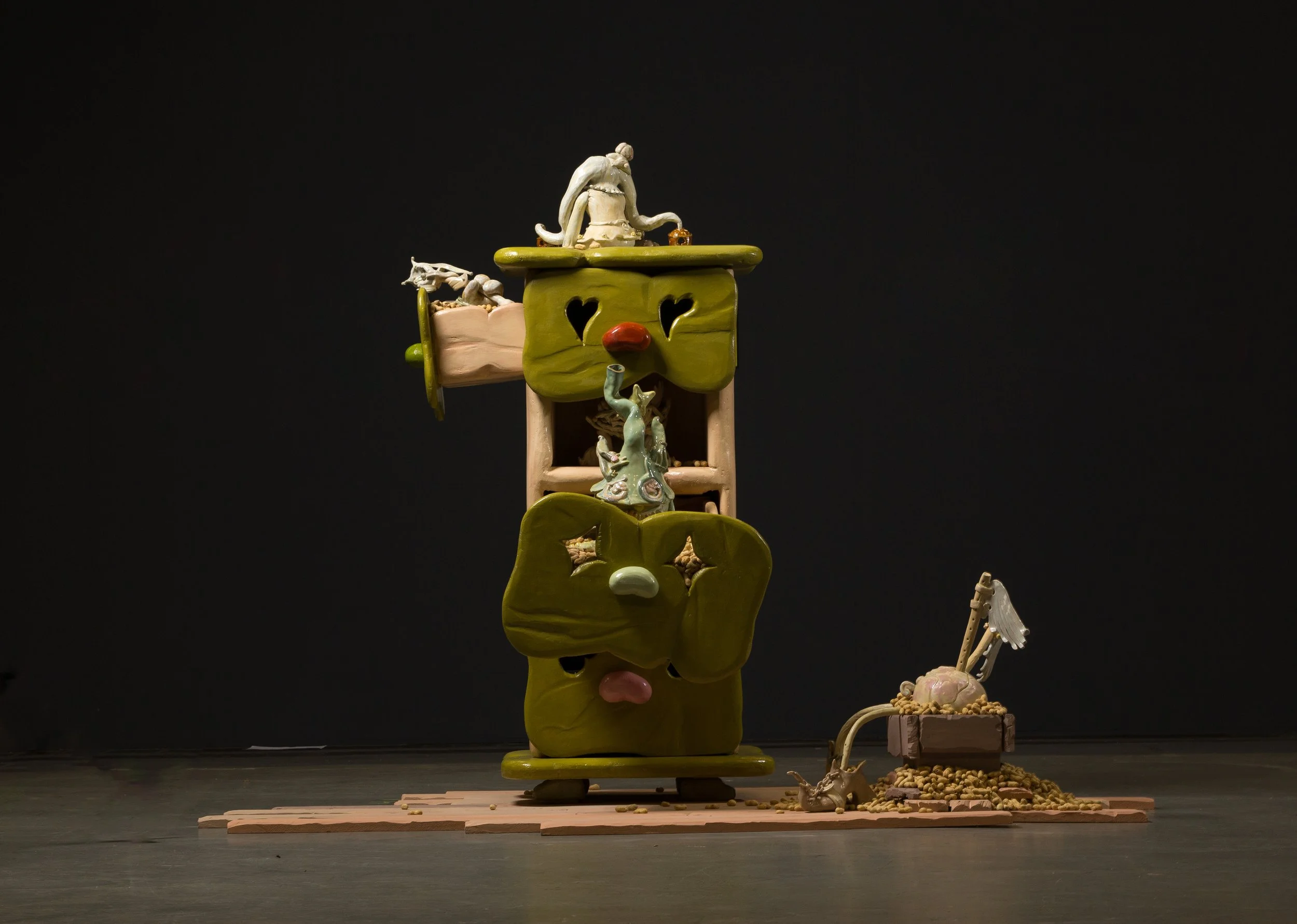 An art installation consisting of ceramic sculptures stacked to resemble a face, with various abstract elements and a small model of a skeleton on a cart nearby, set against a black background.