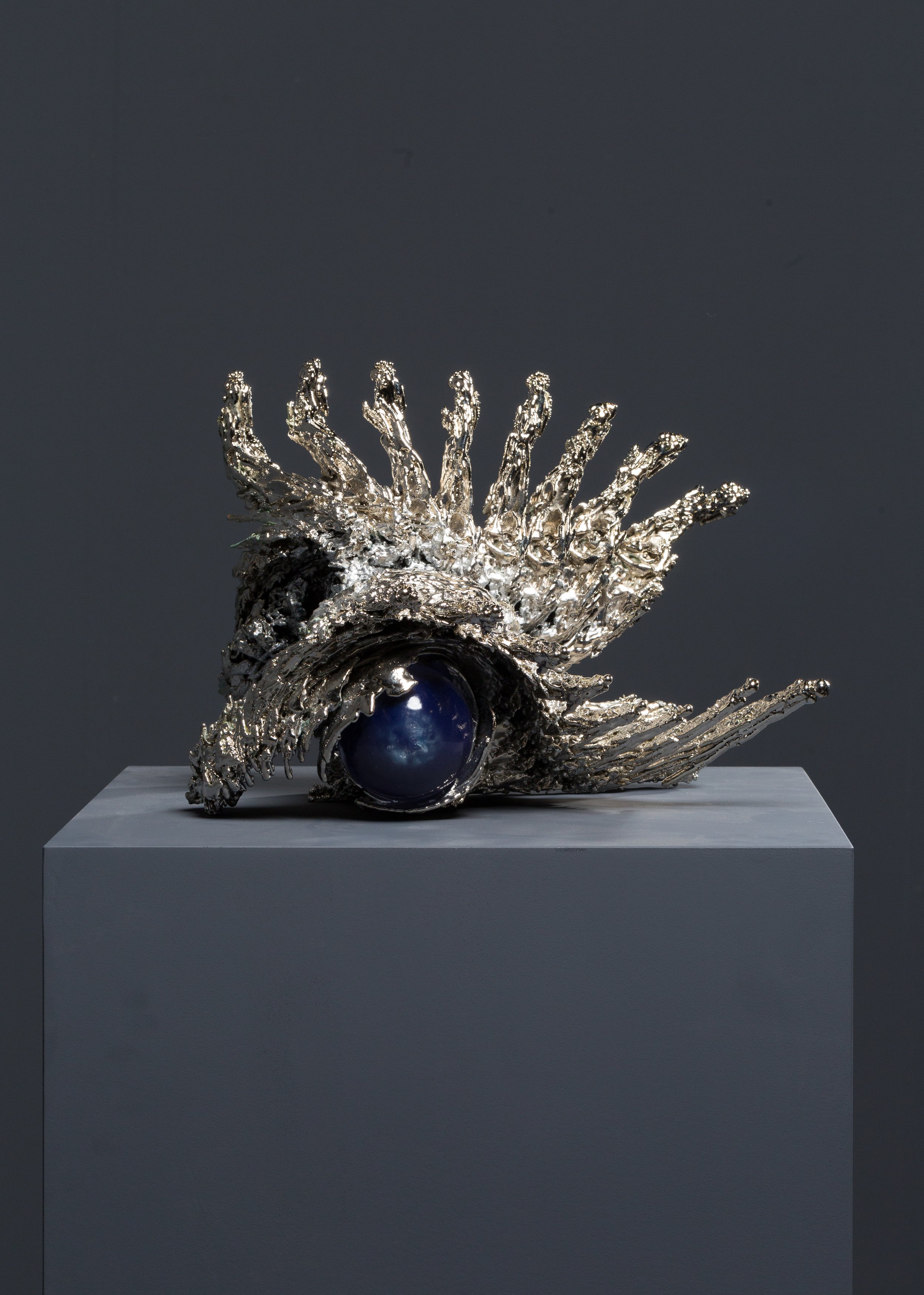 Silver sculpture resembling a hand with elongated, jagged fingers, set on a gray pedestal with a dark gray background.