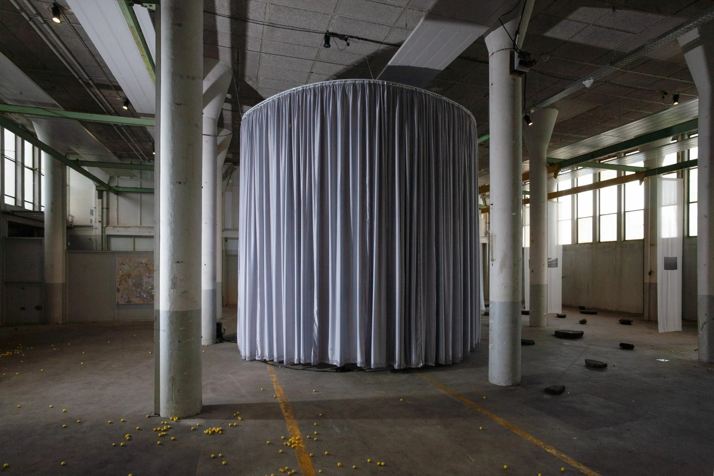 An industrial-style interior with a cluster of white vertical columns and large windows, featuring a cylindrical, metallic, pleated structure suspended in the center, with scattered yellow balls on the floor.