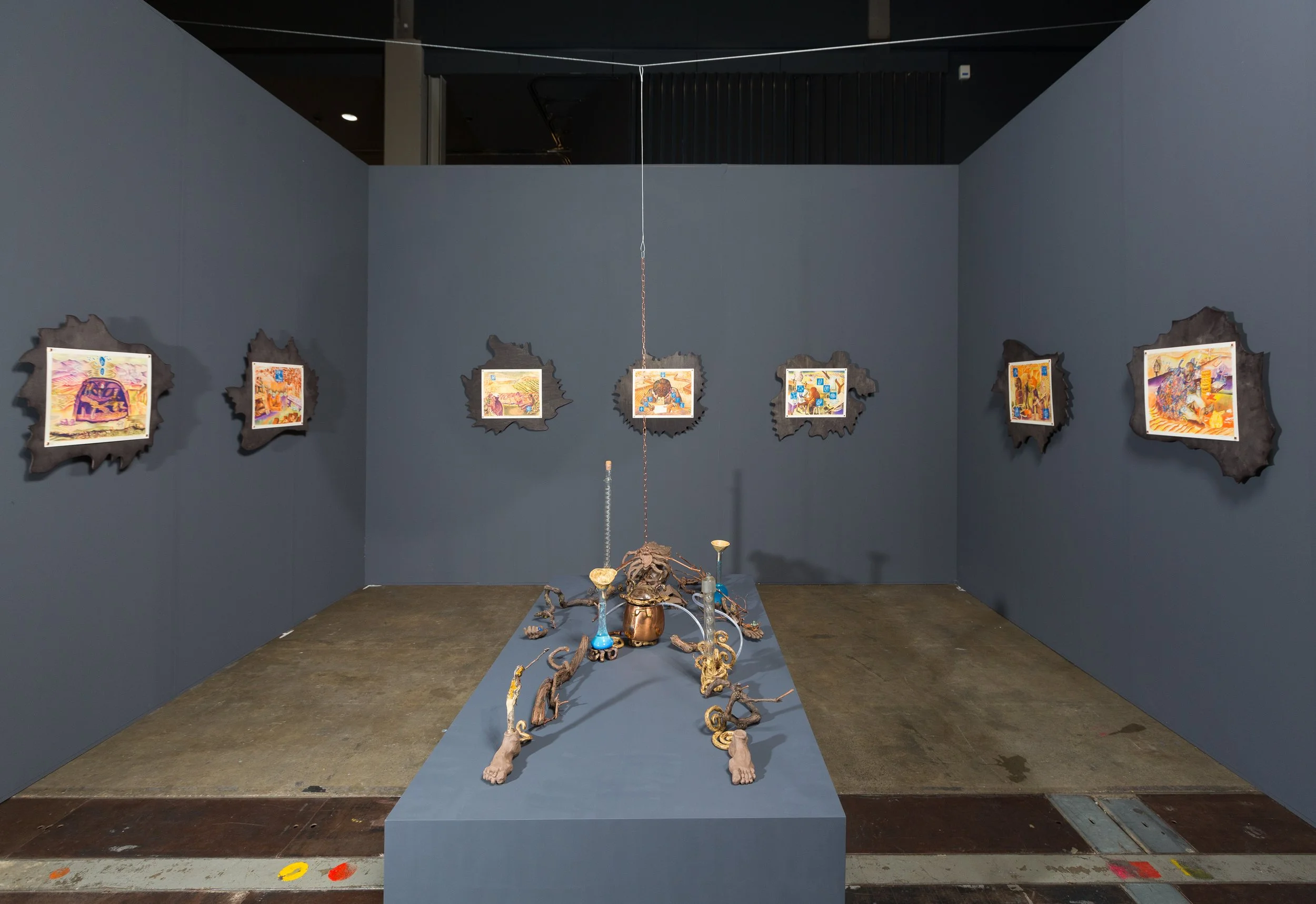 An art gallery display featuring framed illustrations on a dark gray wall and a sculptural piece on a blue platform in the center. The sculpture appears to be a creative assembly of natural and metallic objects, resembling legs and feet with various embellishments.
