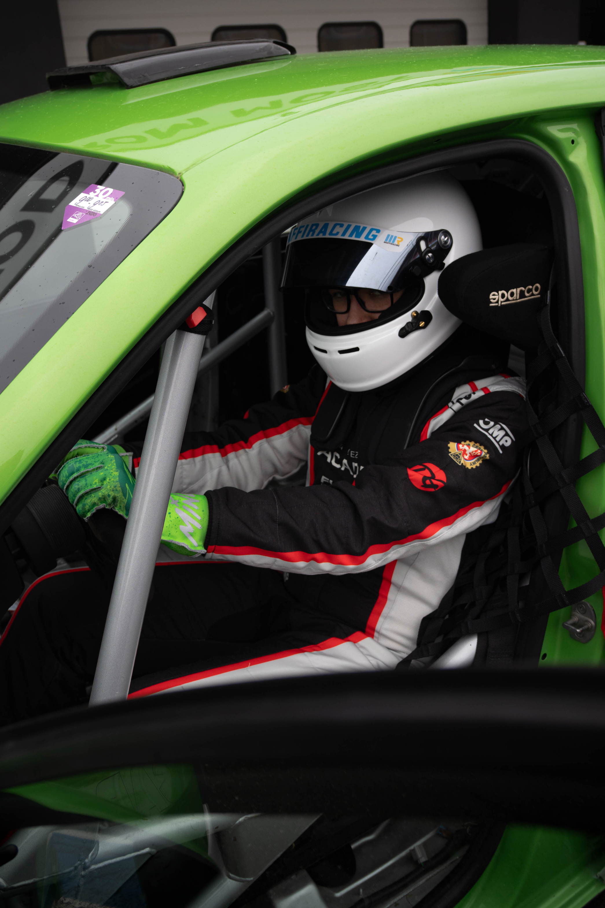 A race car driver wearing a white helmet with blue and yellow details, glasses, and a racing suit with red and black accents, seated inside a bright green race car.