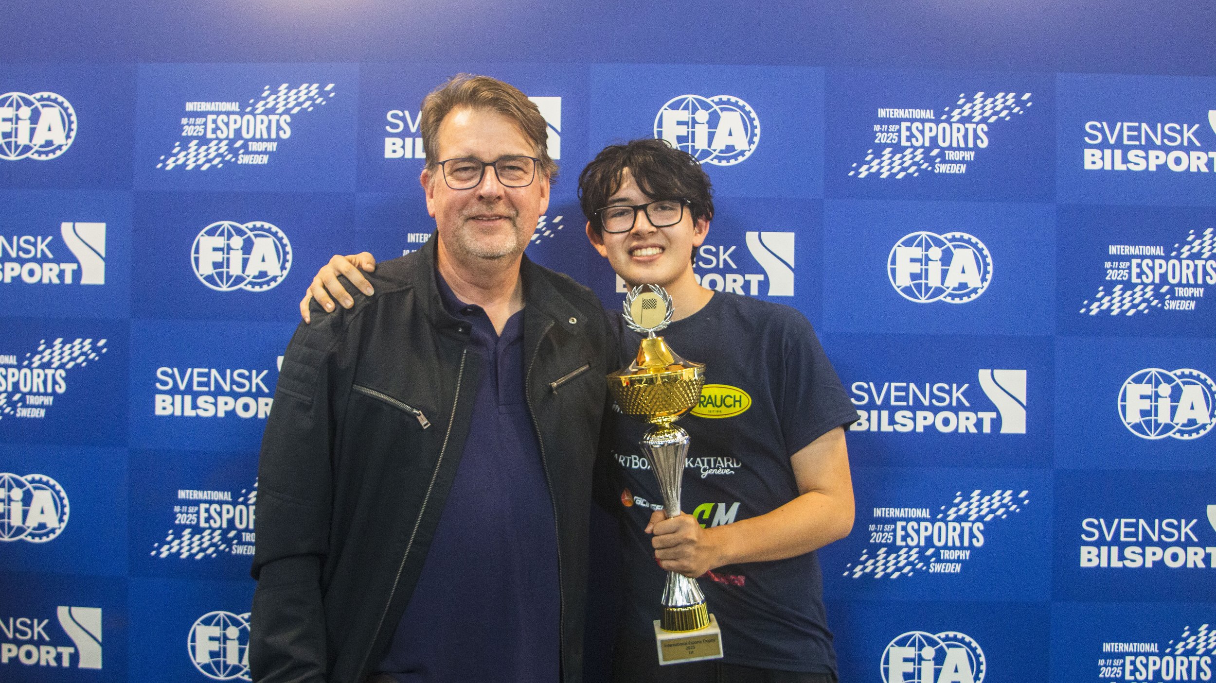A grown man with glasses and a beard standing next to a young woman with glasses, smiling, holding a trophy, at an esports event with a blue backdrop that has various logos and text.
