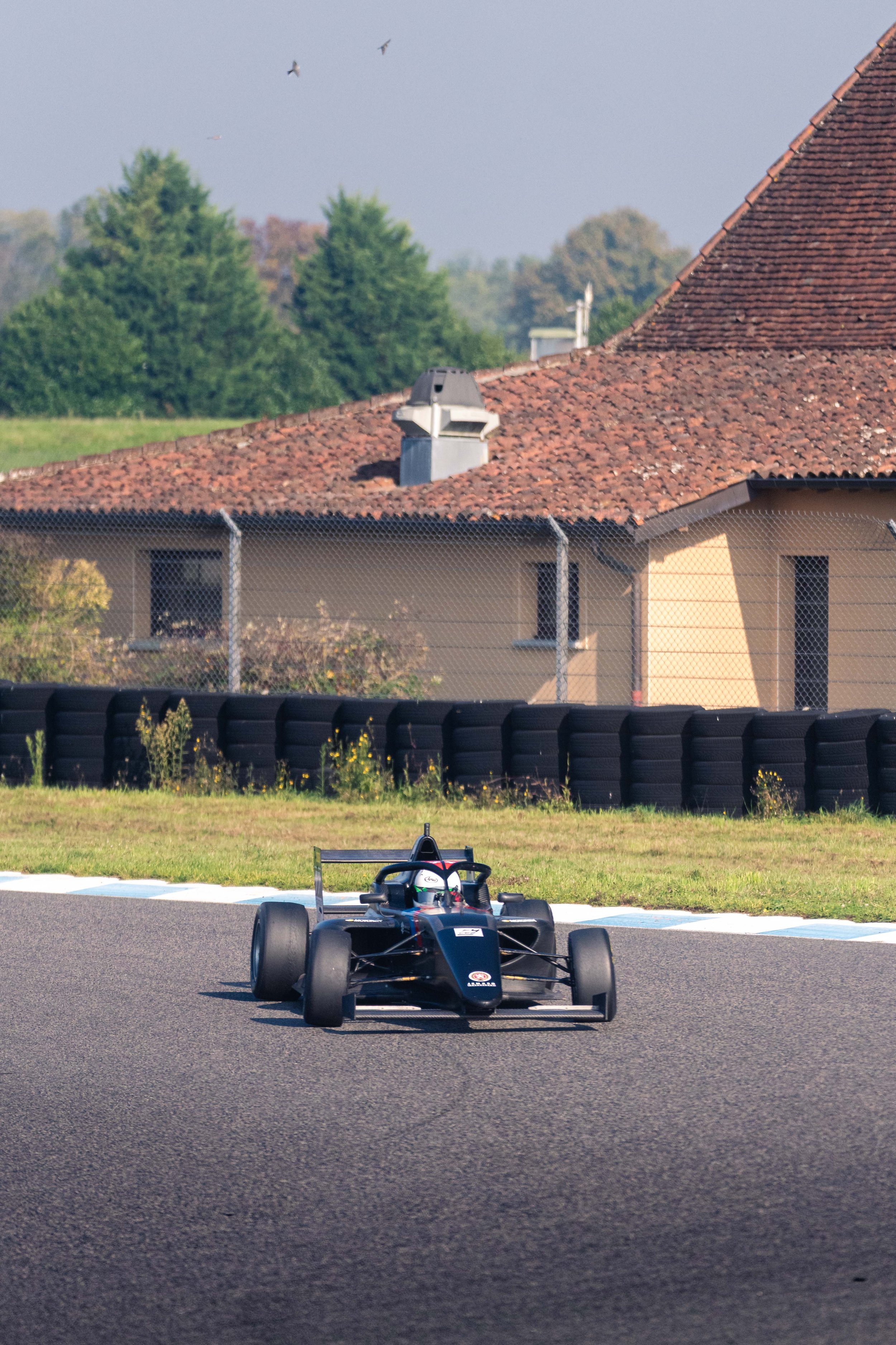 A black race car with open wheels and a high rear wing on a race track, with a house and trees in the background and a chain-link fence separating the track from the surroundings.