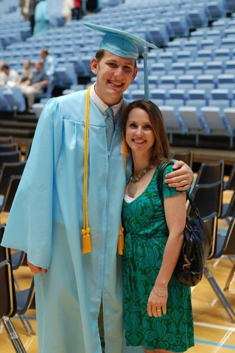 A young man in a blue graduation cap and gown with a gold cord, smiling and standing next to a woman in a green dress, indoors at a graduation ceremony.
