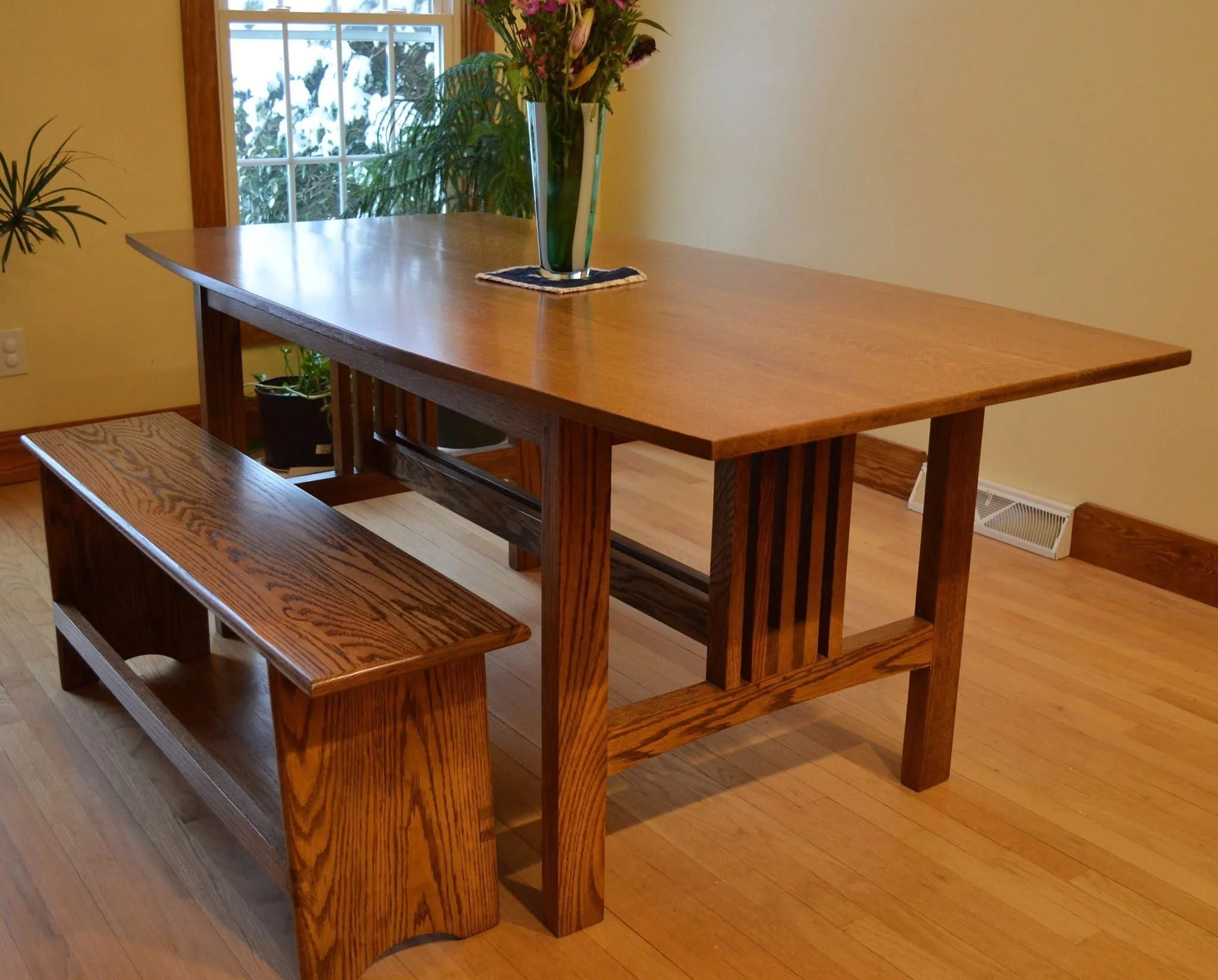 Wooden dining table with a bench on one side and a vase of flowers on top, situated in a room with hardwood flooring and a window with snow outside.