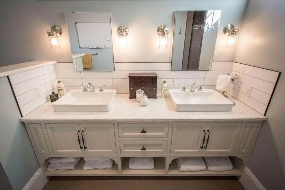 Double bathroom vanity with white sinks, mirrors, wall-mounted lights, and folded towels underneath.