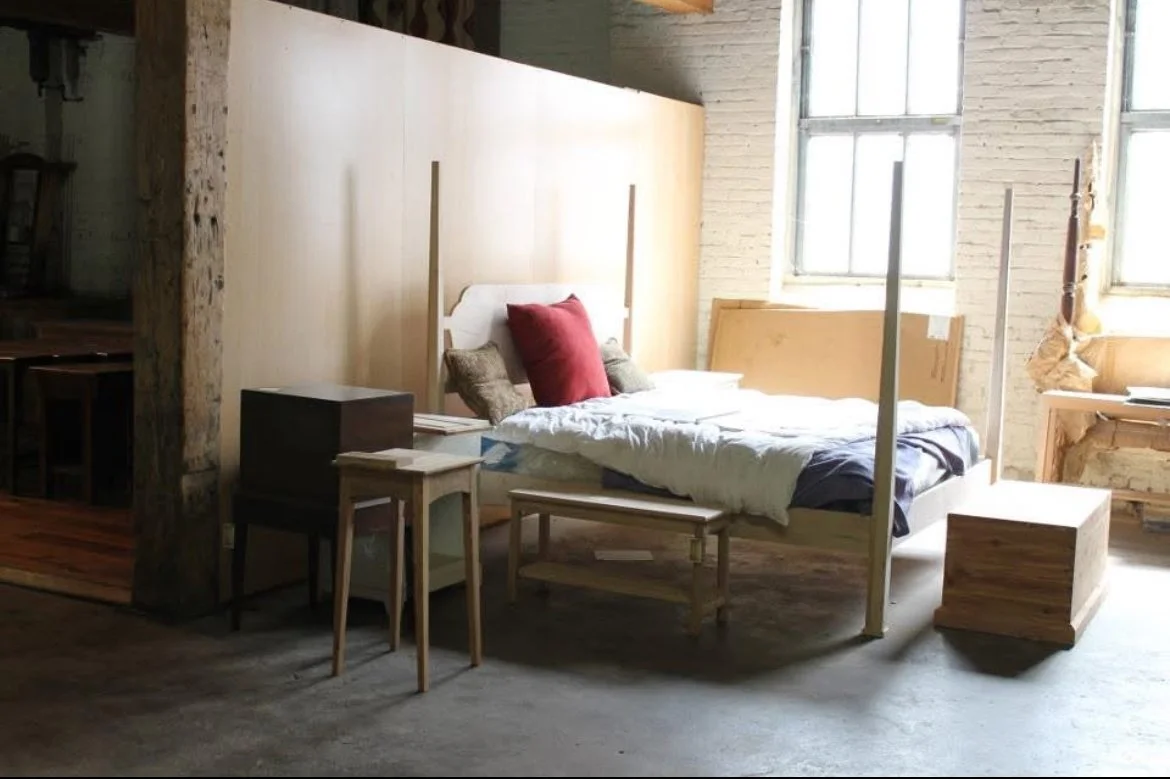 A bedroom with a four-poster bed, pillows, nightstand, and natural light coming through large windows.