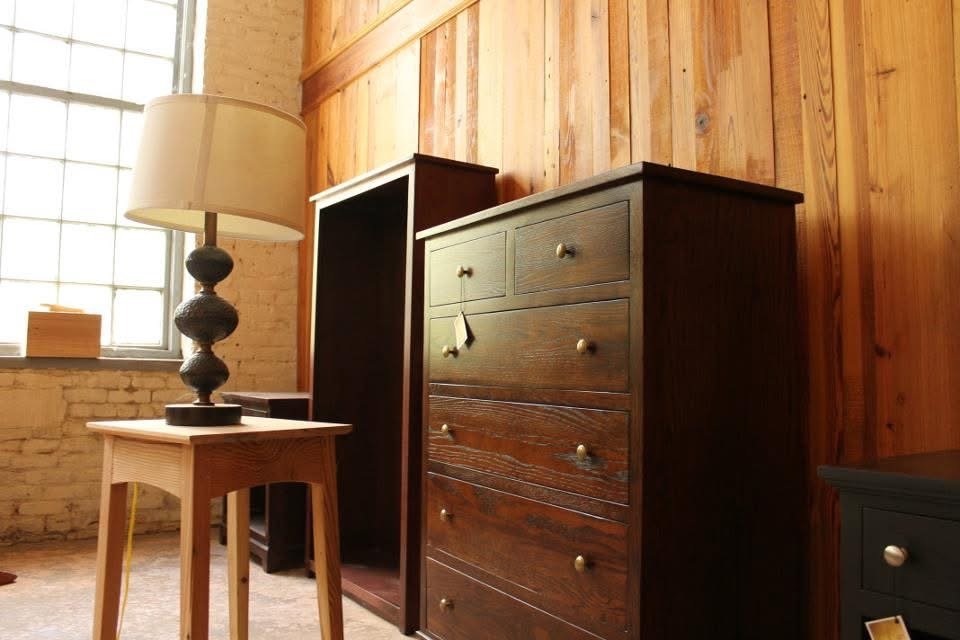 Interior of a room with a large window, wooden walls, a beige table lamp on a small light-colored wooden table, and dark-colored wooden dressers.