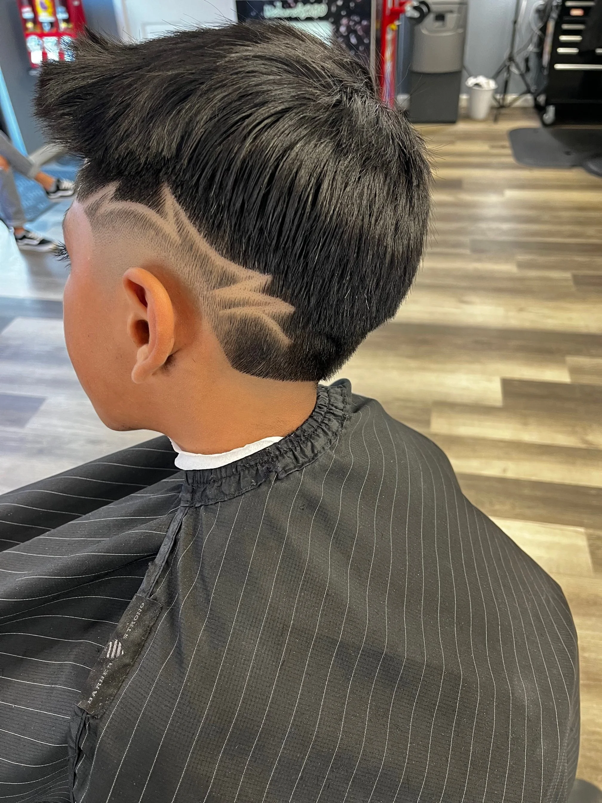 A young man with a modern fade haircut featuring a lightning bolt design shaved into the side of his head, sitting in a barbershop.