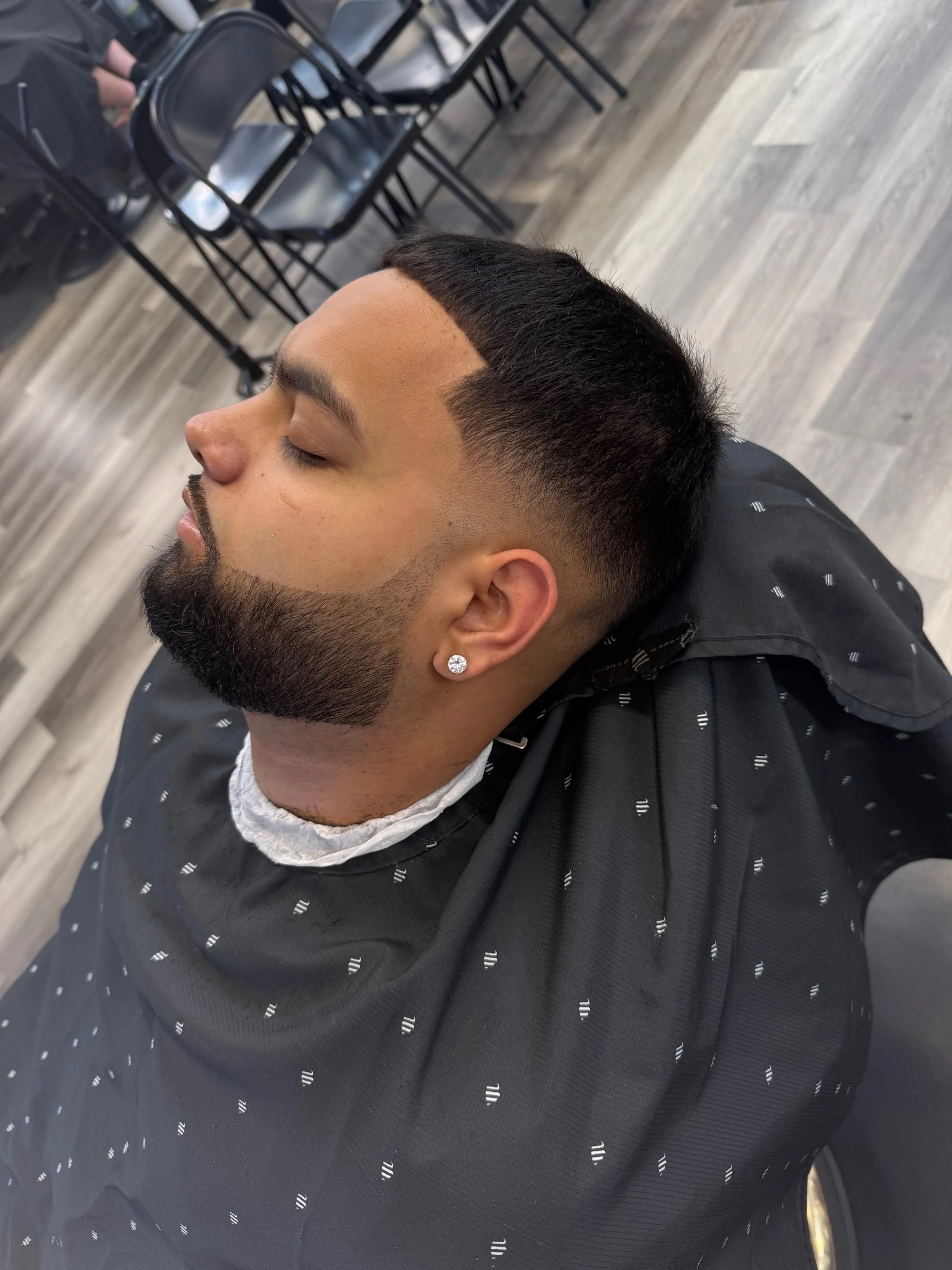 Close-up of a man with a freshly cut hairstyle, featuring a high fade and a sharp, geometric hairline design, sitting in a barber's chair with his eyes closed.
