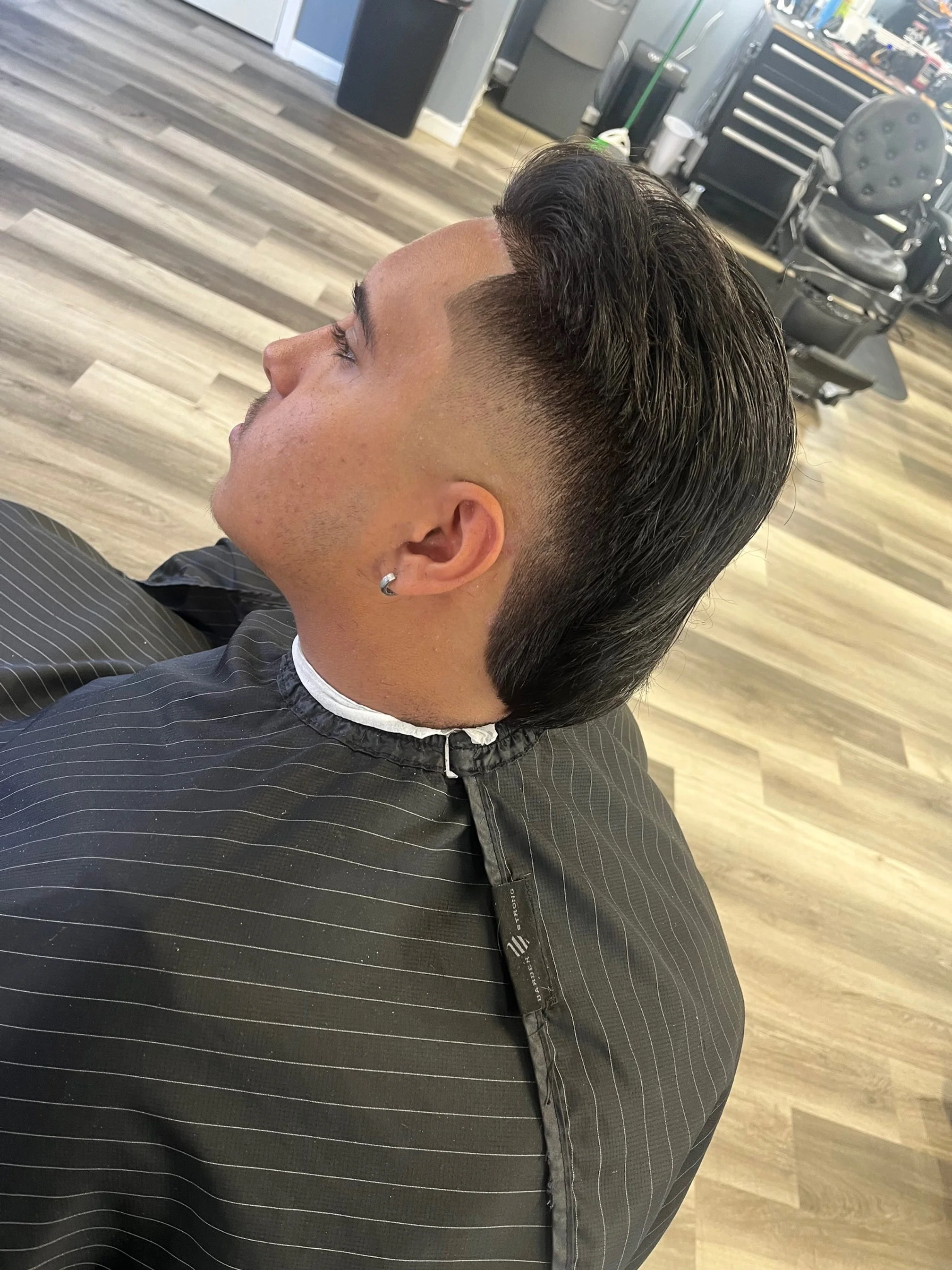 A man with a fresh haircut sitting in a barber shop chair, wearing a black striped cape. The barber shop has wood flooring, barber chairs, and equipment visible in the background.