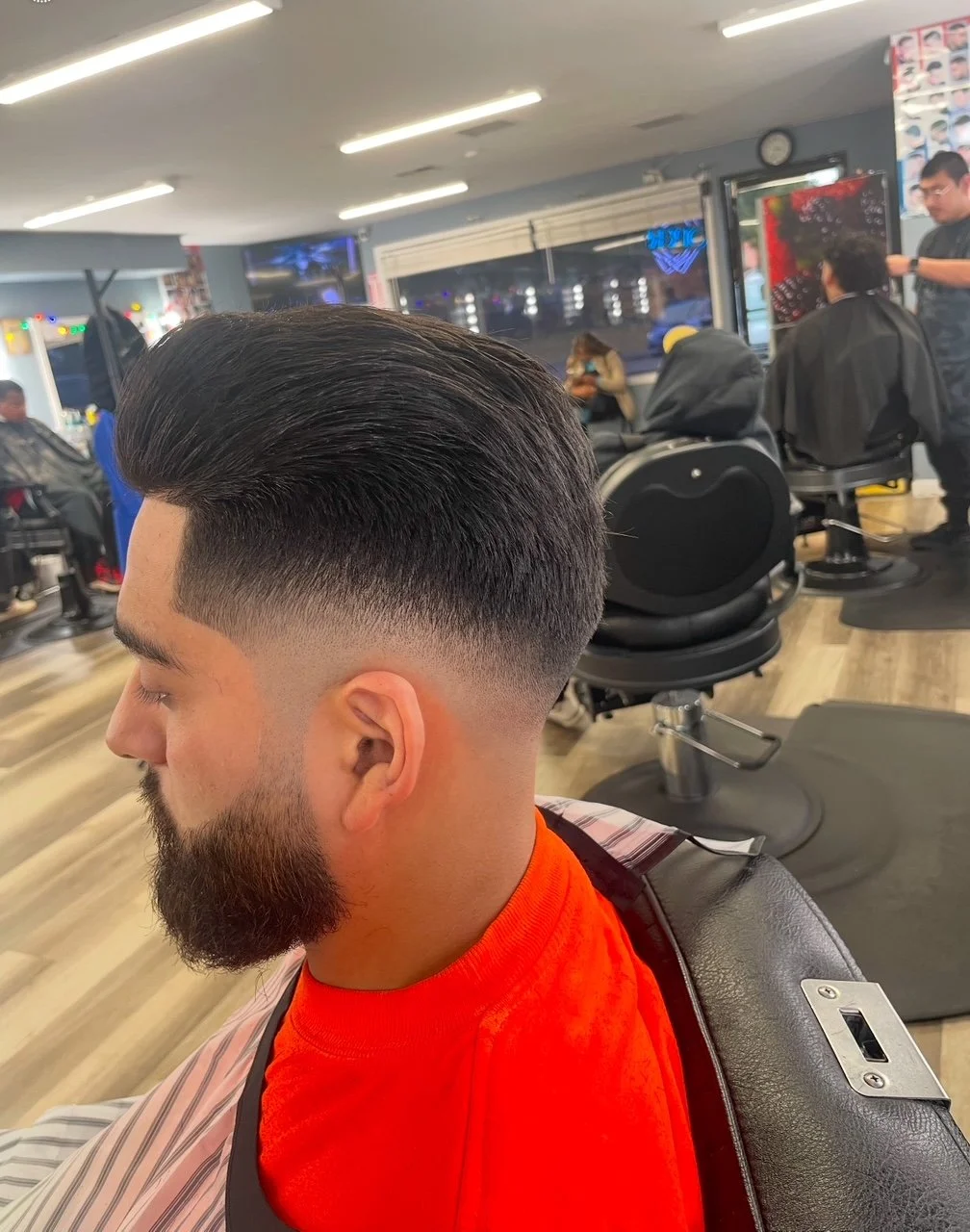 Man with a fresh high fade hairstyle and beard sitting in barbershop chair, barber working on client's hair in background, barbershop interior with chairs, mirrors, and customers.