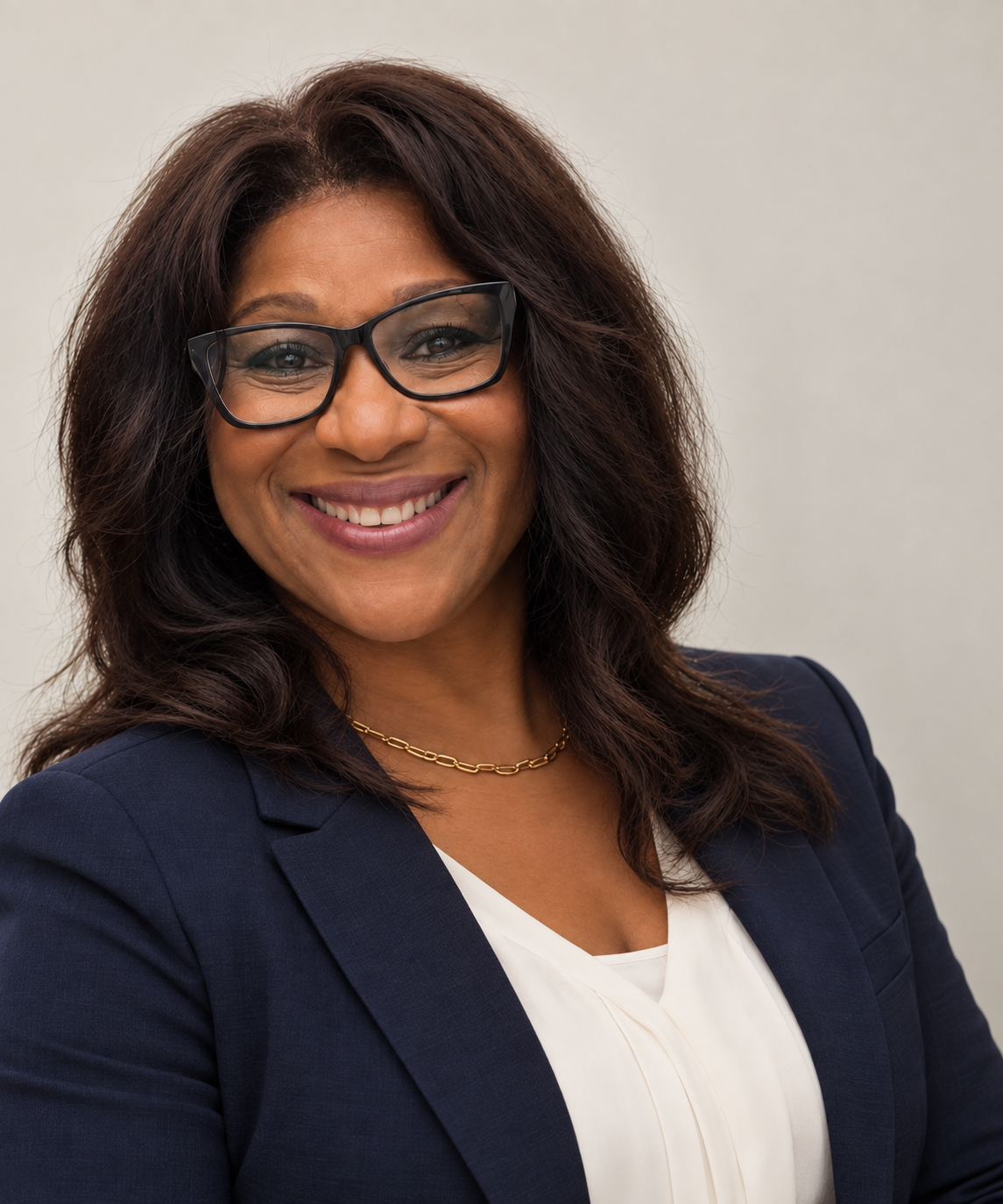 A woman with brown hair wearing glasses, a navy blazer, and a gold chain necklace, smiling in front of a plain light-colored background.