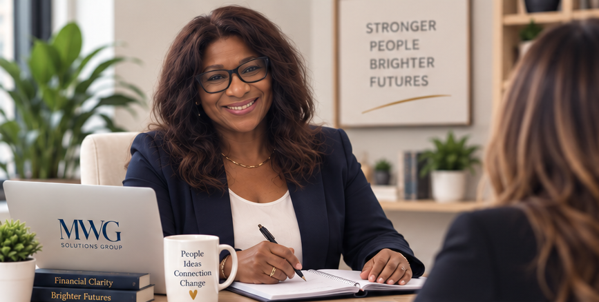 A woman with glasses and curly hair smiling during a meeting in an office, with a notebook, a mug, and books on the desk.