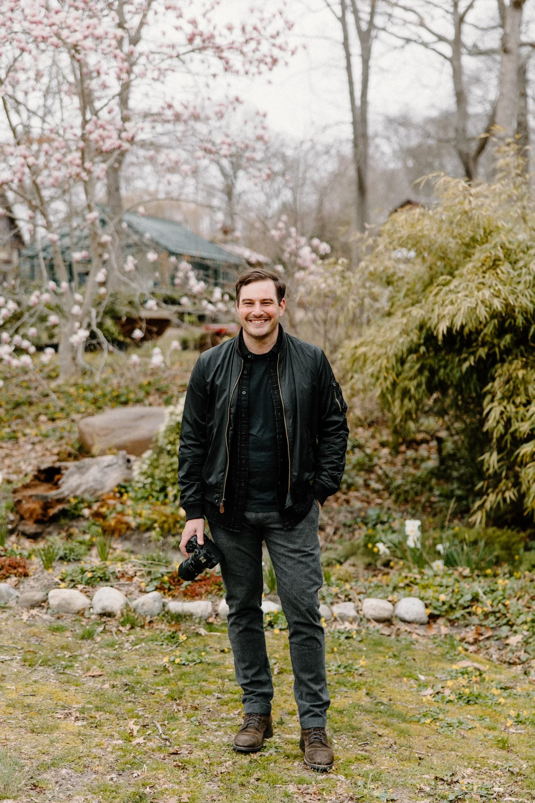 A smiling man with a camera in hand standing outdoors in a garden with blooming trees and shrubs, overcast sky.