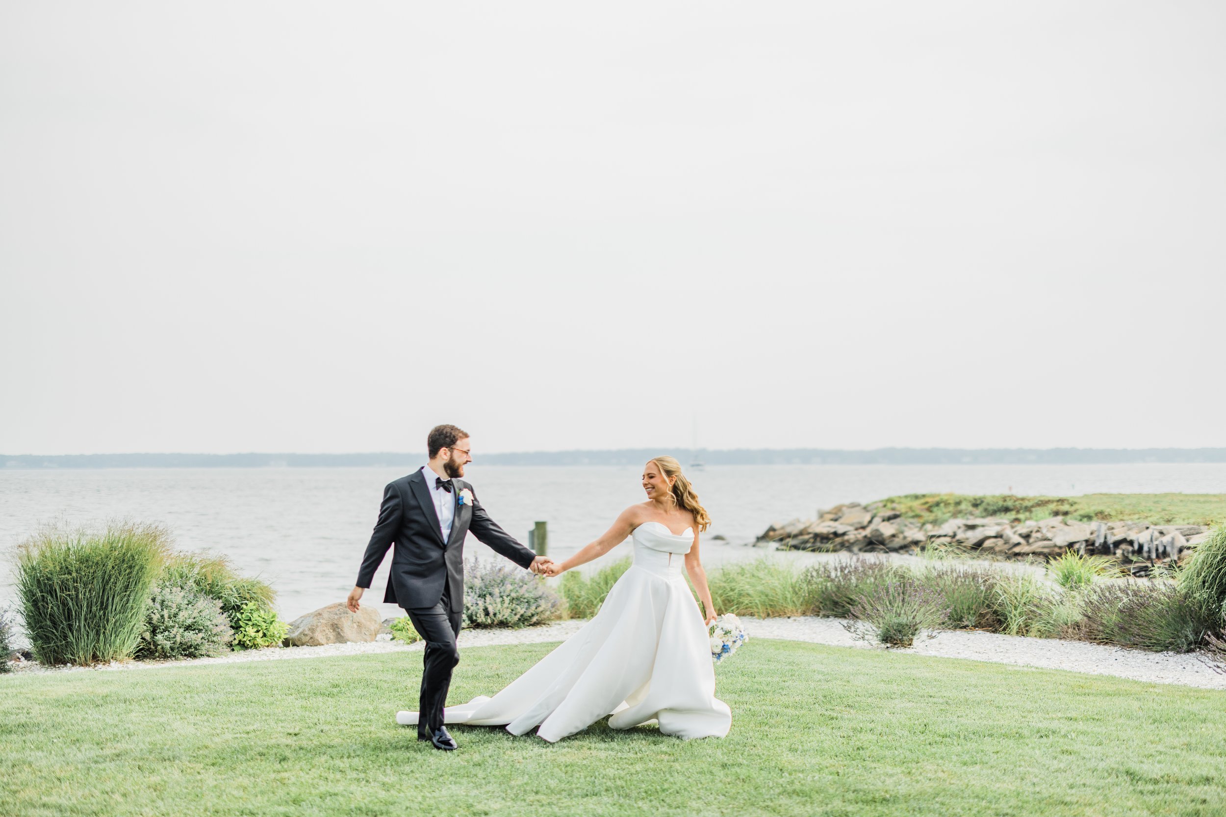 A bride and groom holding hands outdoors near a body of water, with grass and rocks in the background on a cloudy day.