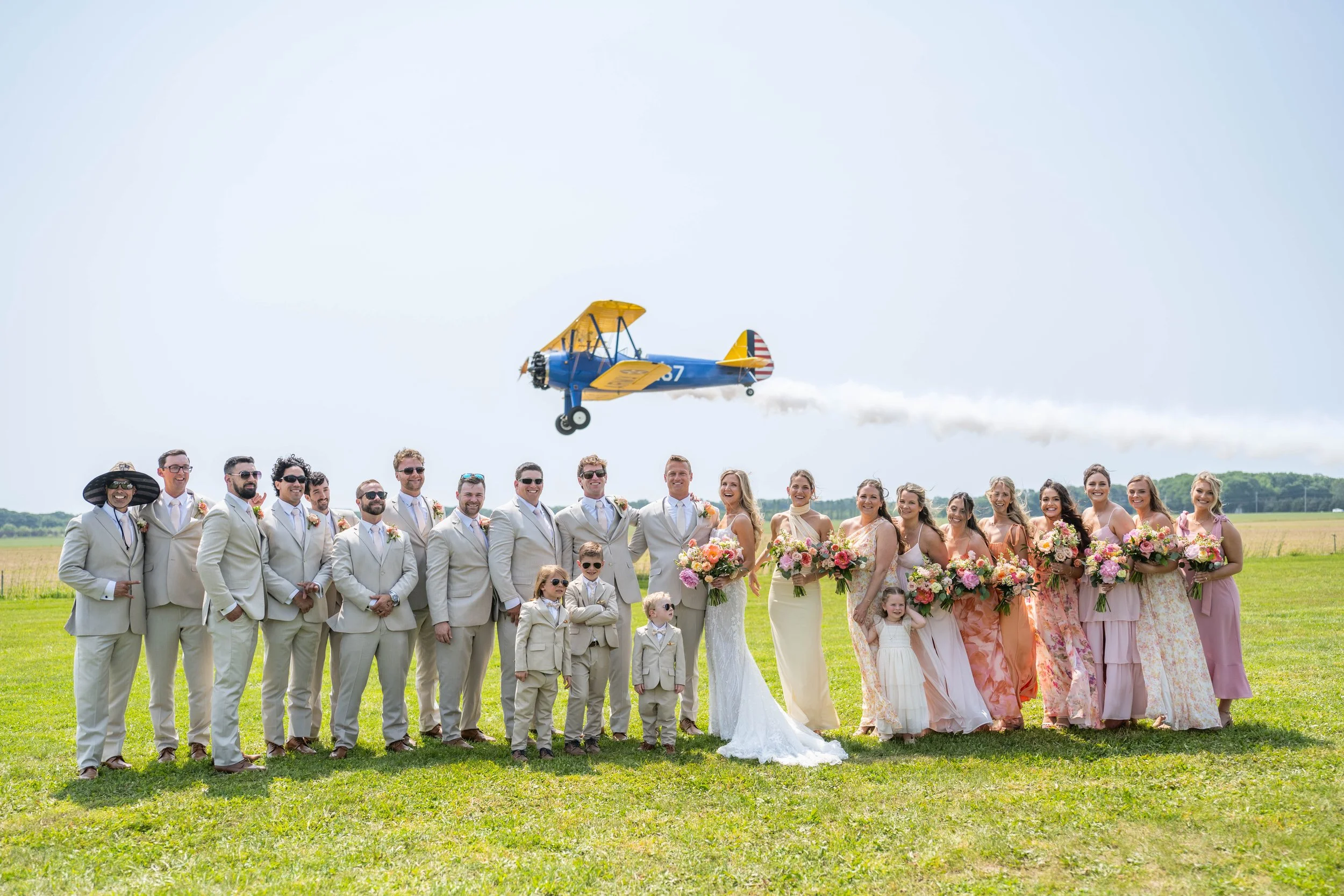 A wedding party standing on a grassy field with a blue and yellow airplane flying overhead, emitting smoke.