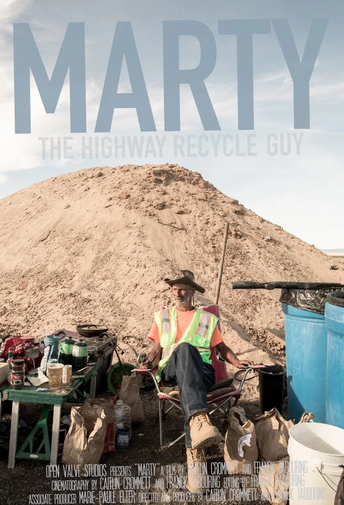 A man wearing a safety vest and a wide-brimmed hat sitting outdoors with a large dirt mound behind him. He has a beard and is smiling, sitting in a folding chair near a table with various containers and supplies. There are large blue plastic barrels to his right and paper bags around him.