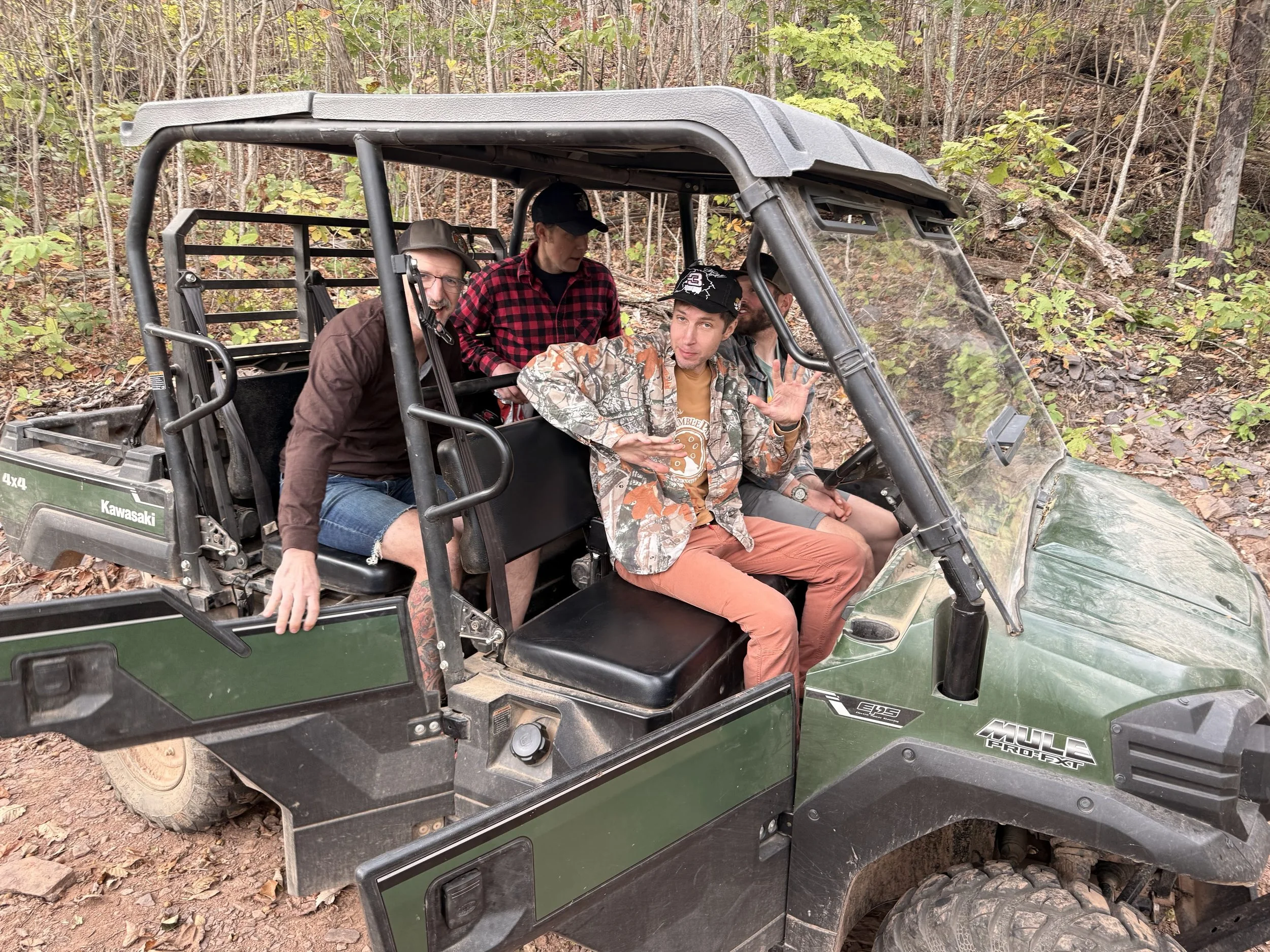 Four men sitting and standing inside a green off-road utility vehicle in a wooded area with fallen leaves and trees in the background.