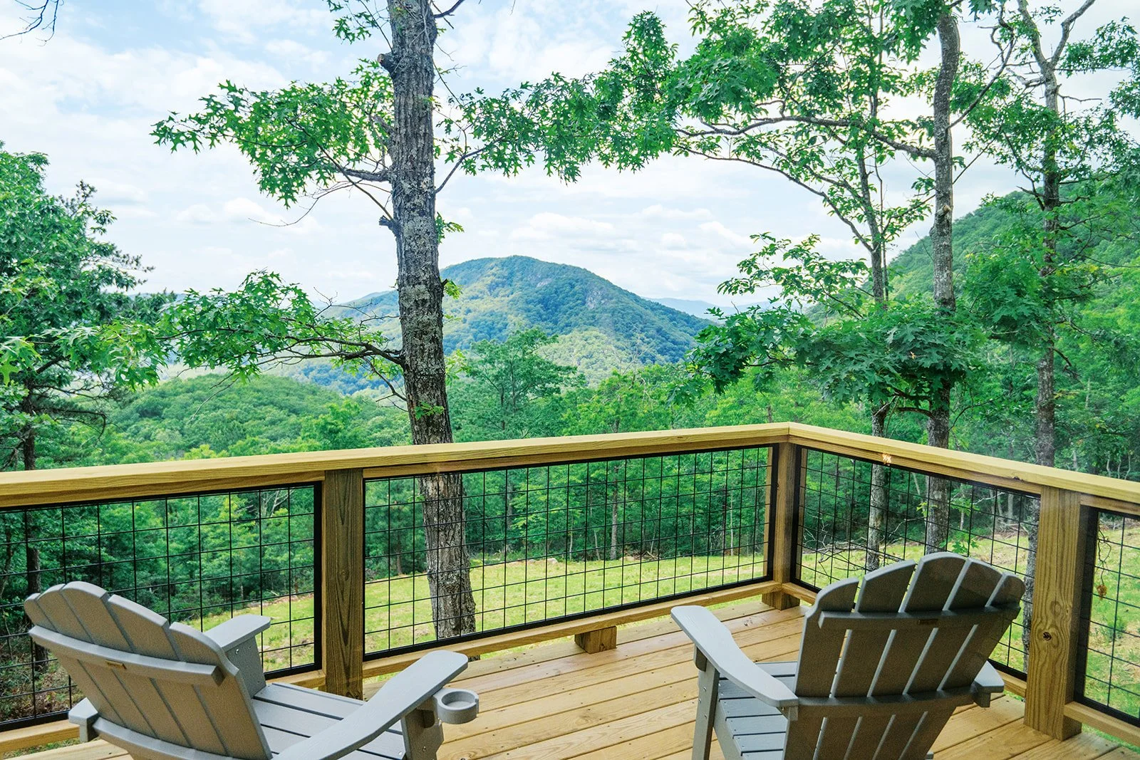 A wooden balcony with two Adirondack chairs overlooking a lush green forest and distant mountains under a partly cloudy sky.