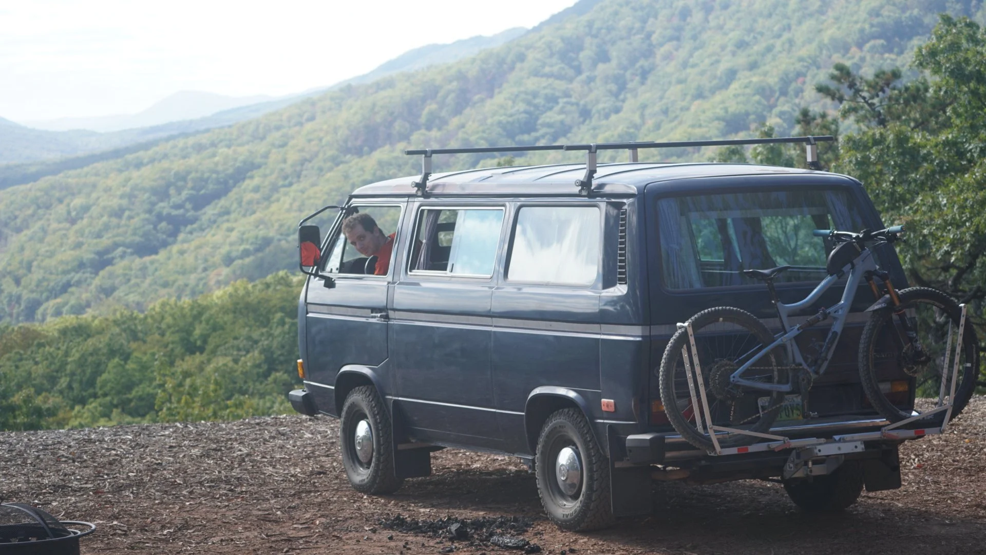 A blue van with a bike rack on the back holding a mountain bike, parked on a dirt trail with green mountains in the background, and a person inside looking out the window.