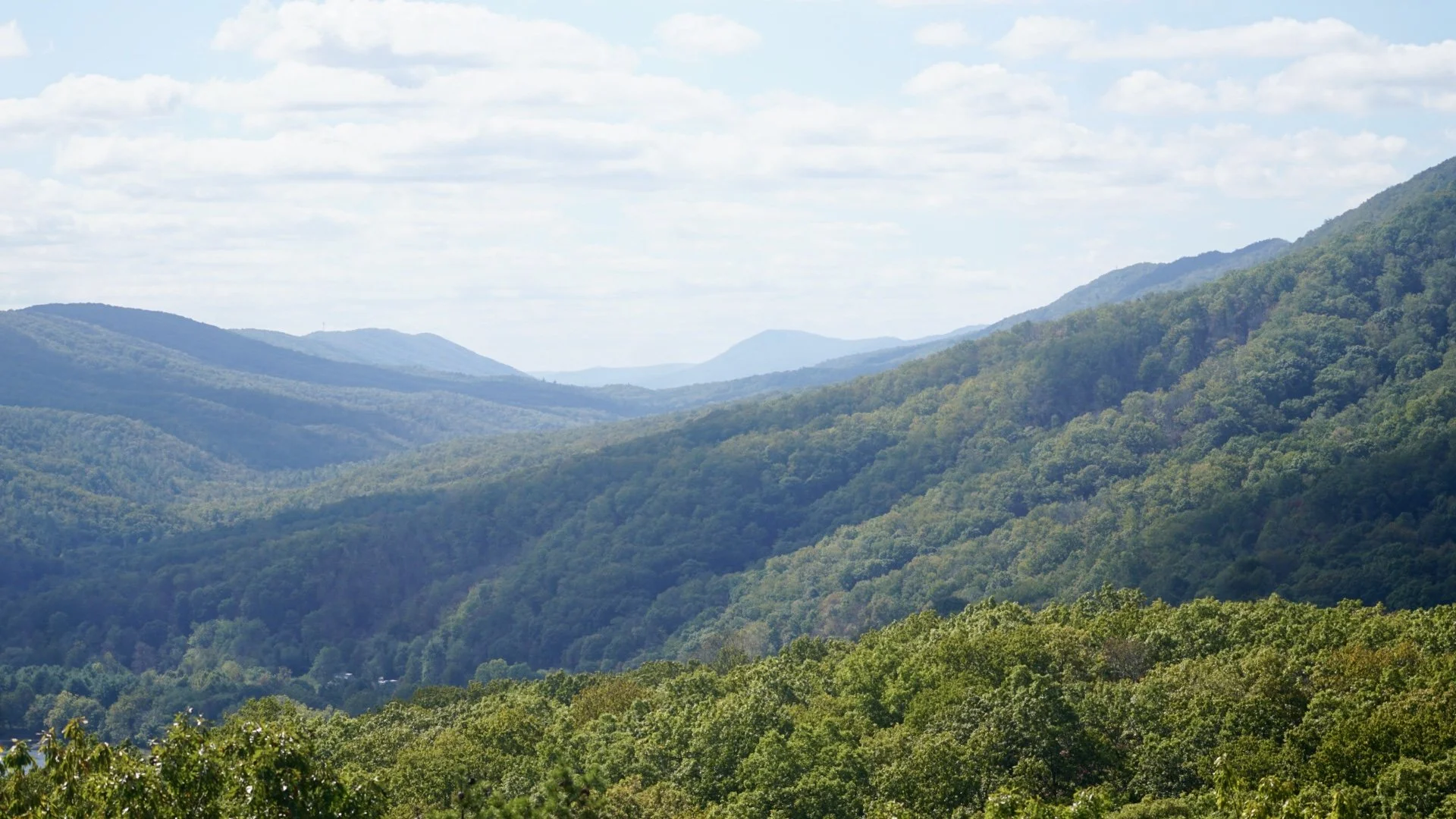 A expansive mountain landscape with multiple ridges covered in green forests, under a partly cloudy sky.