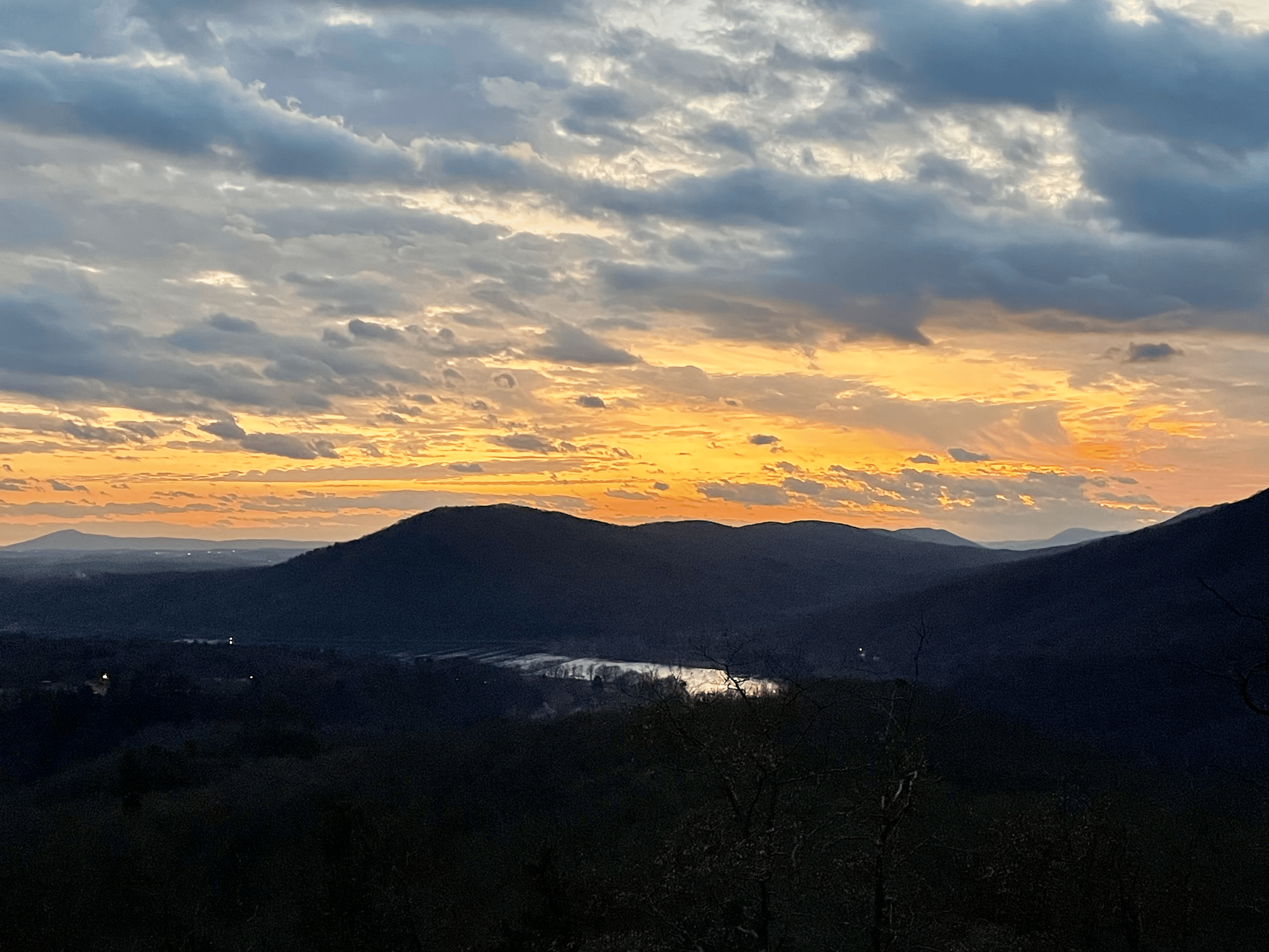 A scenic view of a mountain landscape during sunset with orange and yellow hues in the sky, scattered clouds, and dark silhouettes of mountains and trees in the foreground.