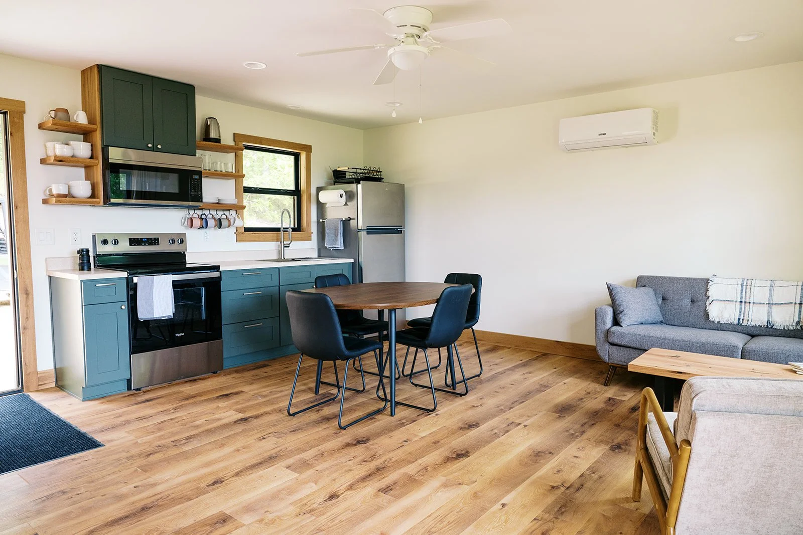 Open-concept living room and kitchen with hardwood floors, teal kitchen cabinets, stainless steel appliances, black dining chairs around a wooden table, gray sofa with a blanket, and a white wall-mounted air conditioner.