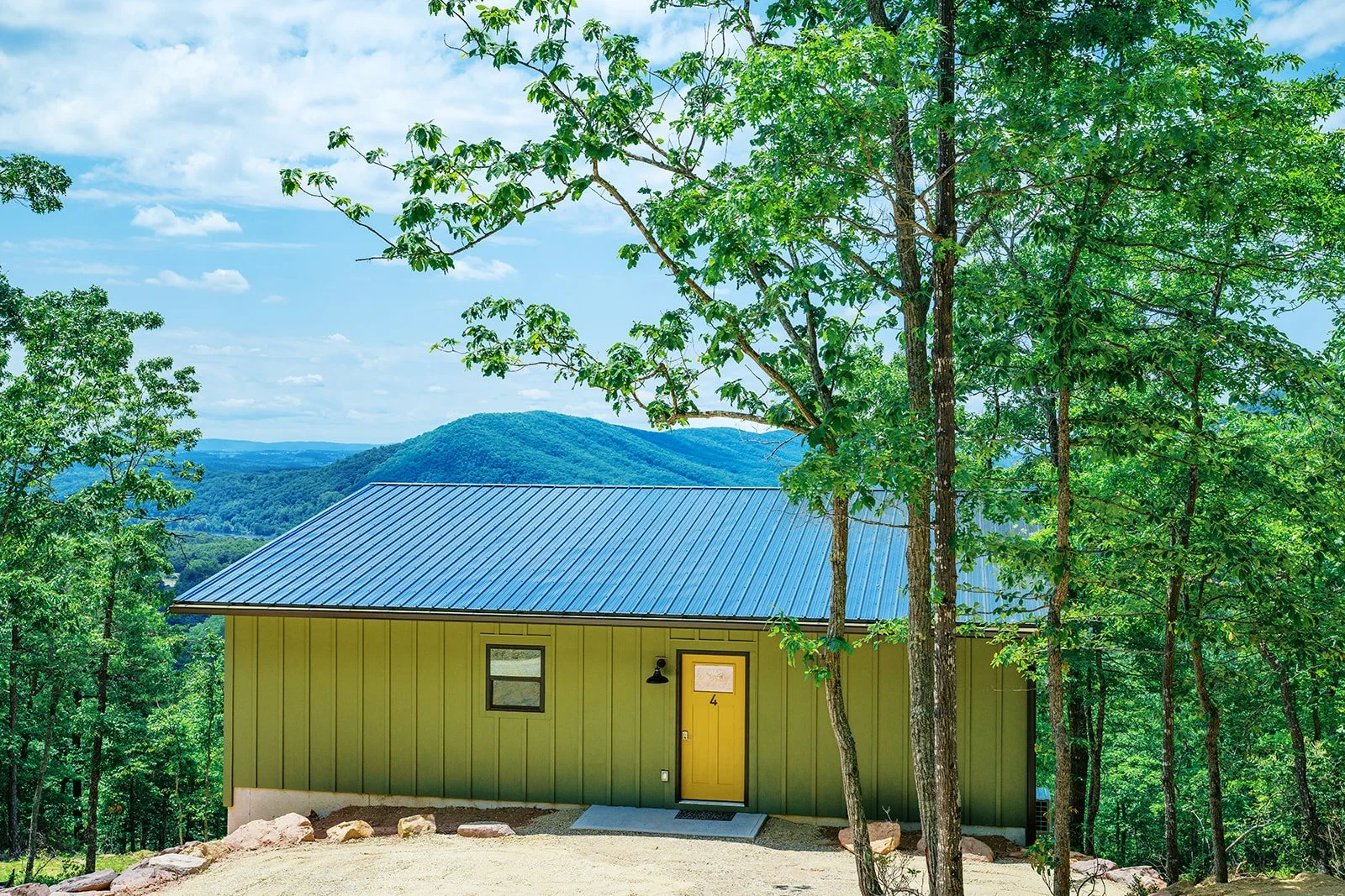 Small yellow house with a green metal roof surrounded by green trees and distant mountains in the background.