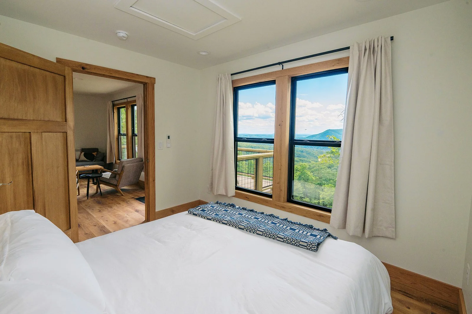 Room with large window overlooking mountains and forest, white curtains, white bed, and wooden accents.