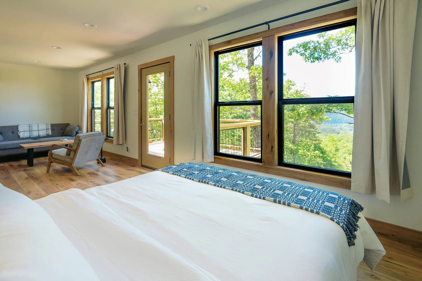 A bedroom with large windows showing green trees outside, a bed with a blue and white blanket, and a sitting area with a sofa, chair, and small table on a wooden floor.