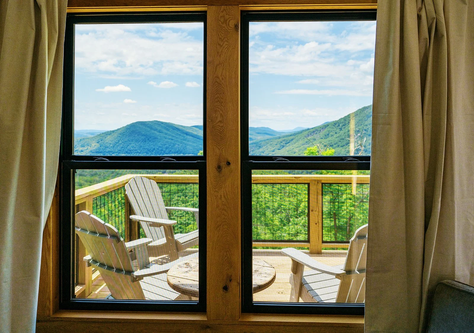 View of a mountain landscape with blue sky and scattered clouds, seen through a window with curtains, leading to a wooden balcony with two Adirondack chairs and a small table.