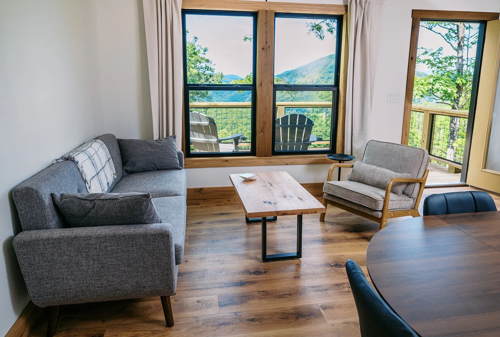 Living room with a gray sofa, a wooden coffee table, a beige armchair, and large windows showing a view of trees and mountains outside.
