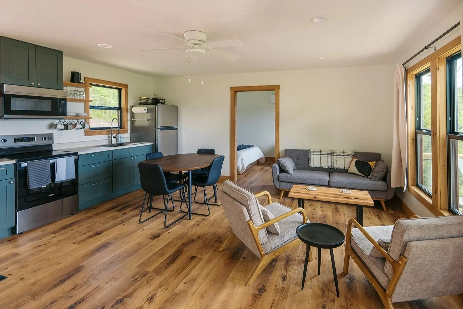 Open-plan living space with a kitchen, dining area, and sitting area. The kitchen has teal cabinets, a microwave, and a refrigerator. The sitting area includes a gray couch, beige armchairs, and a small wooden coffee table. The room has hardwood floors, large windows with wooden frames, and a ceiling fan.
