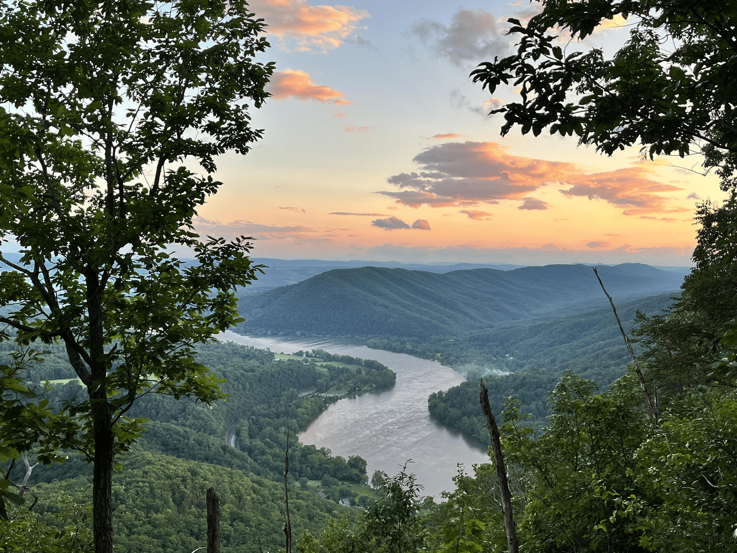 Scenic view of a river flowing through a lush green valley with rolling hills in the background during sunset, framed by tree branches and leaves.