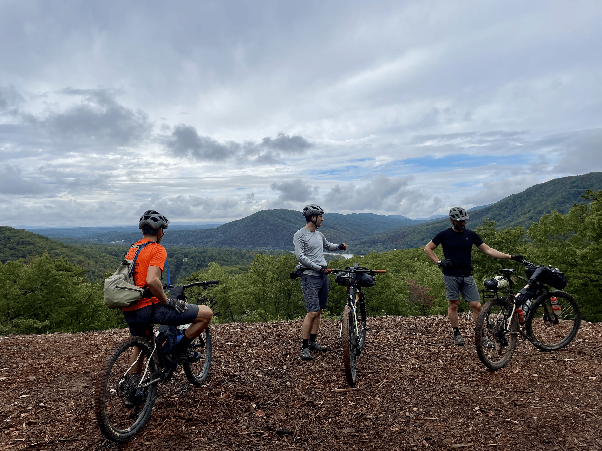 Four men in cycling gear with helmets and backpacks standing with bicycles on a dirt trail overlooking a scenic mountainous landscape under a cloudy sky.
