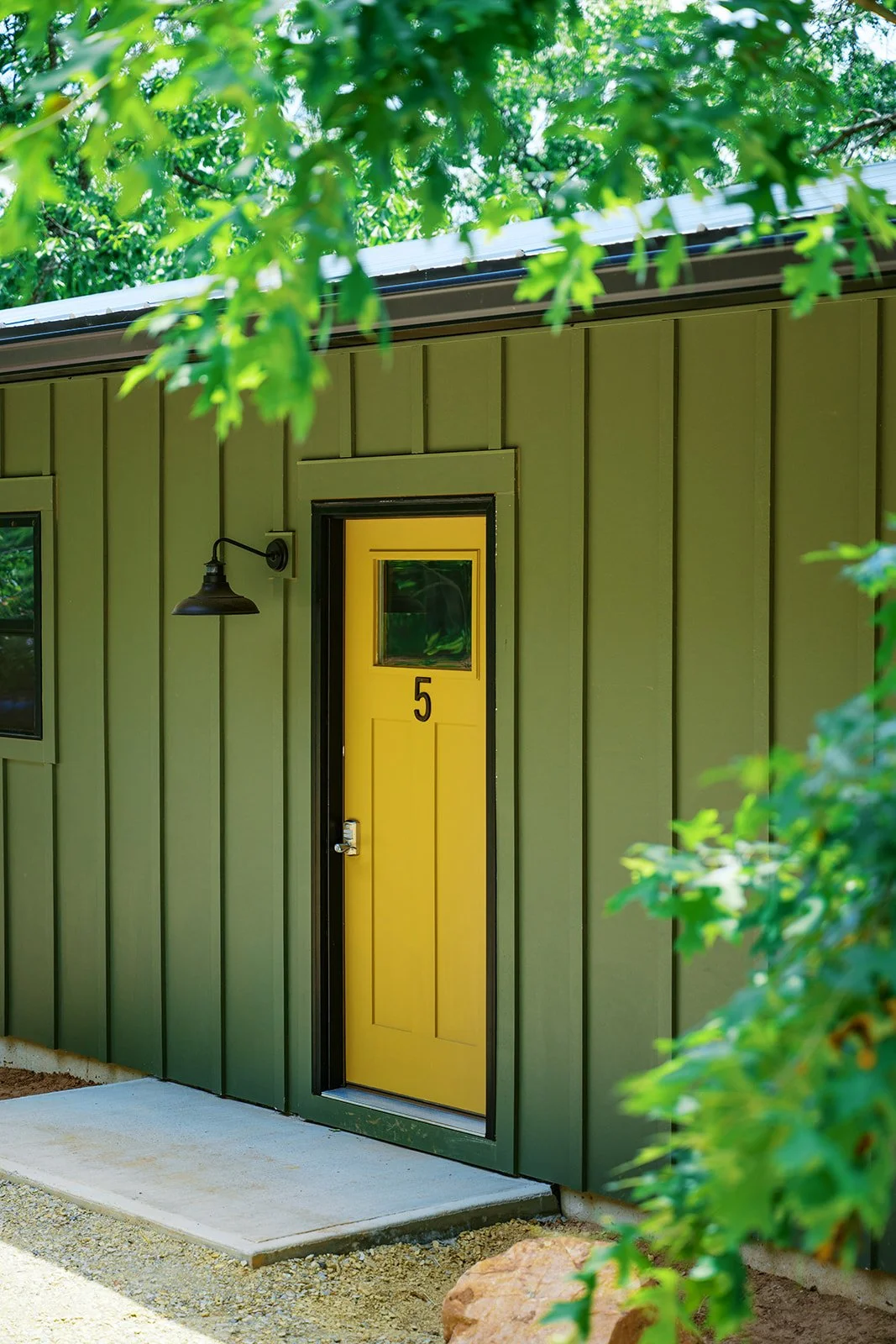 Bright yellow front door with the number 5, situated on a green wall of a house, with a concrete step outside and greenery surrounding the house.