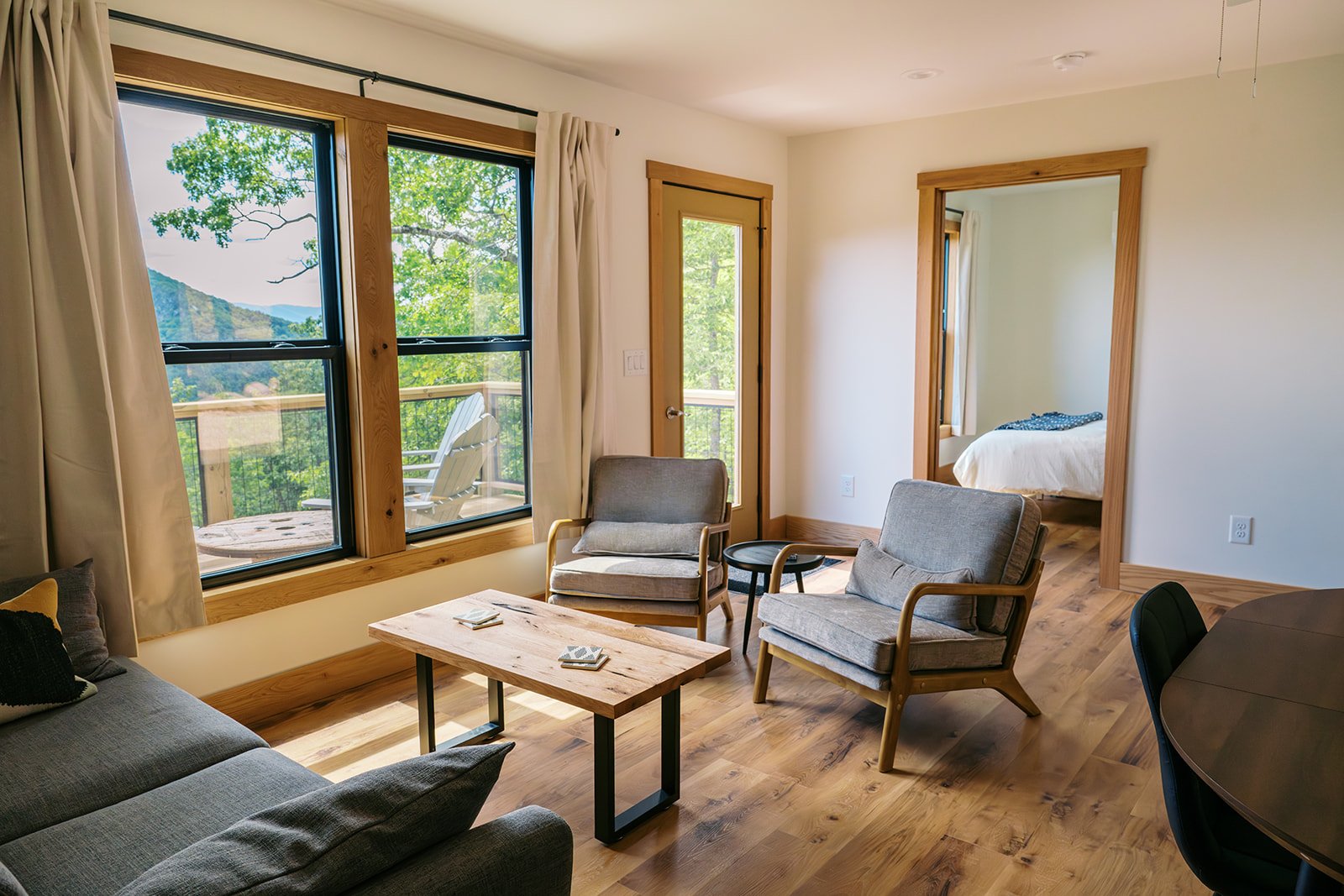 Living room with large windows revealing outdoor trees and mountains, wooden floors, two gray armchairs, a gray sofa, a wooden coffee table with cards, and a partial view of a bedroom through an open door.