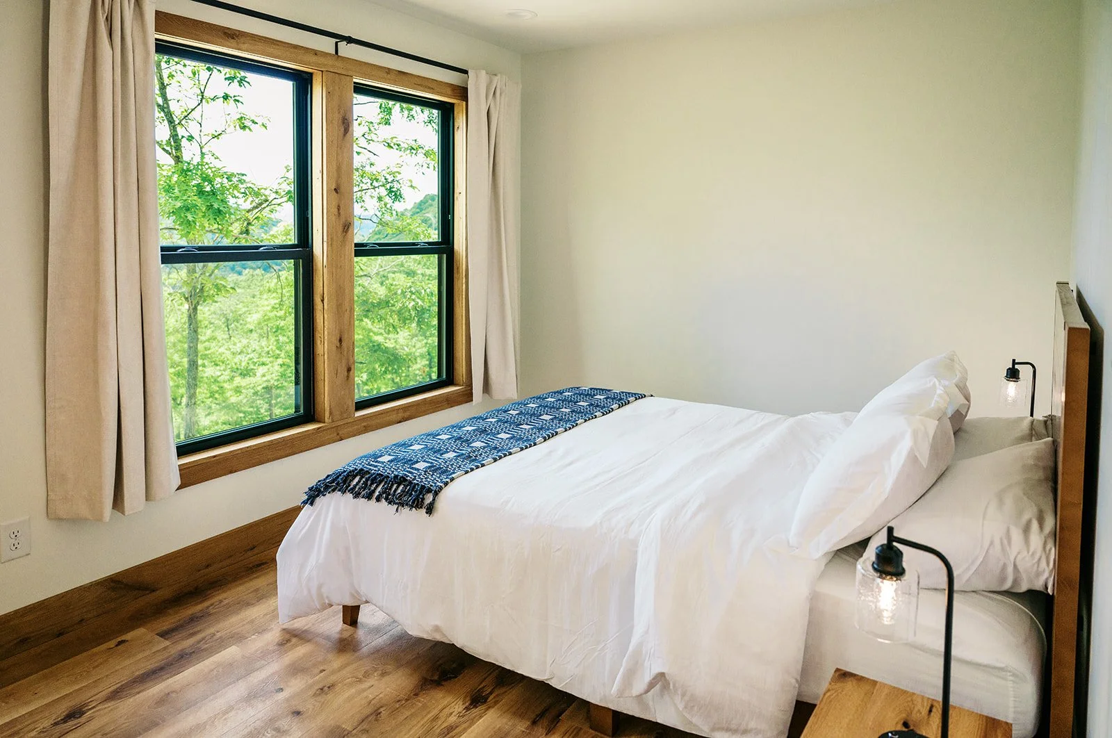 A cozy bedroom with a large window showing green trees outside, white bedding on the bed, beige curtains, and rustic wooden accents.