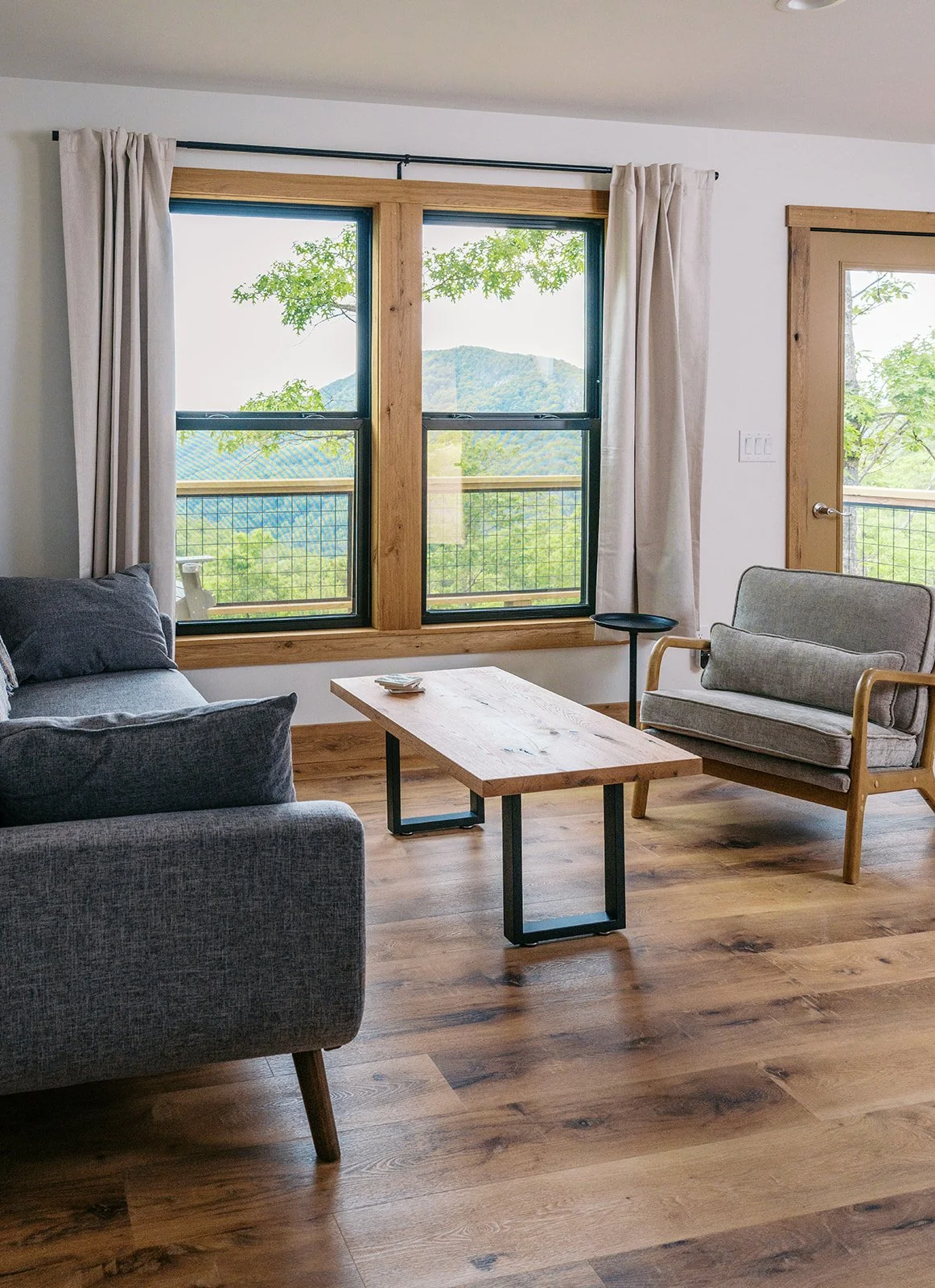 Living room with a large window showing a view of green trees and distant mountains, a gray sofa, a wooden coffee table, an armchair, and a door with a glass panel, all with wooden trim.