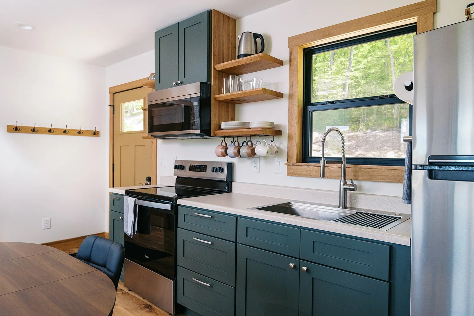 Kitchen with green cabinets, a stove, microwave, open shelves with glasses and plates, a stainless steel refrigerator, a window showing greenery outside, and a wooden dining table with black chairs.