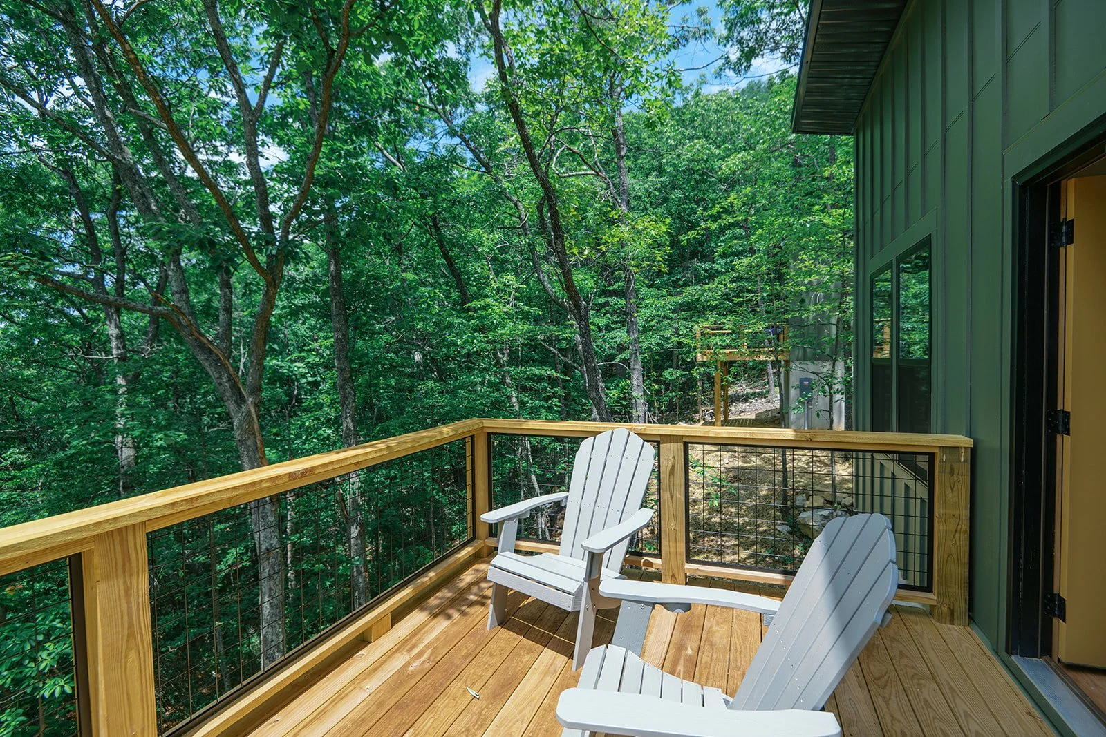 Wooden balcony with two white Adirondack chairs overlooking a dense green forest.