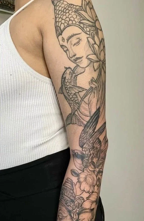A woman with a detailed tattoo sleeve on her arm, featuring a serene face of a woman, two hummingbirds, and various flowers and leaves.